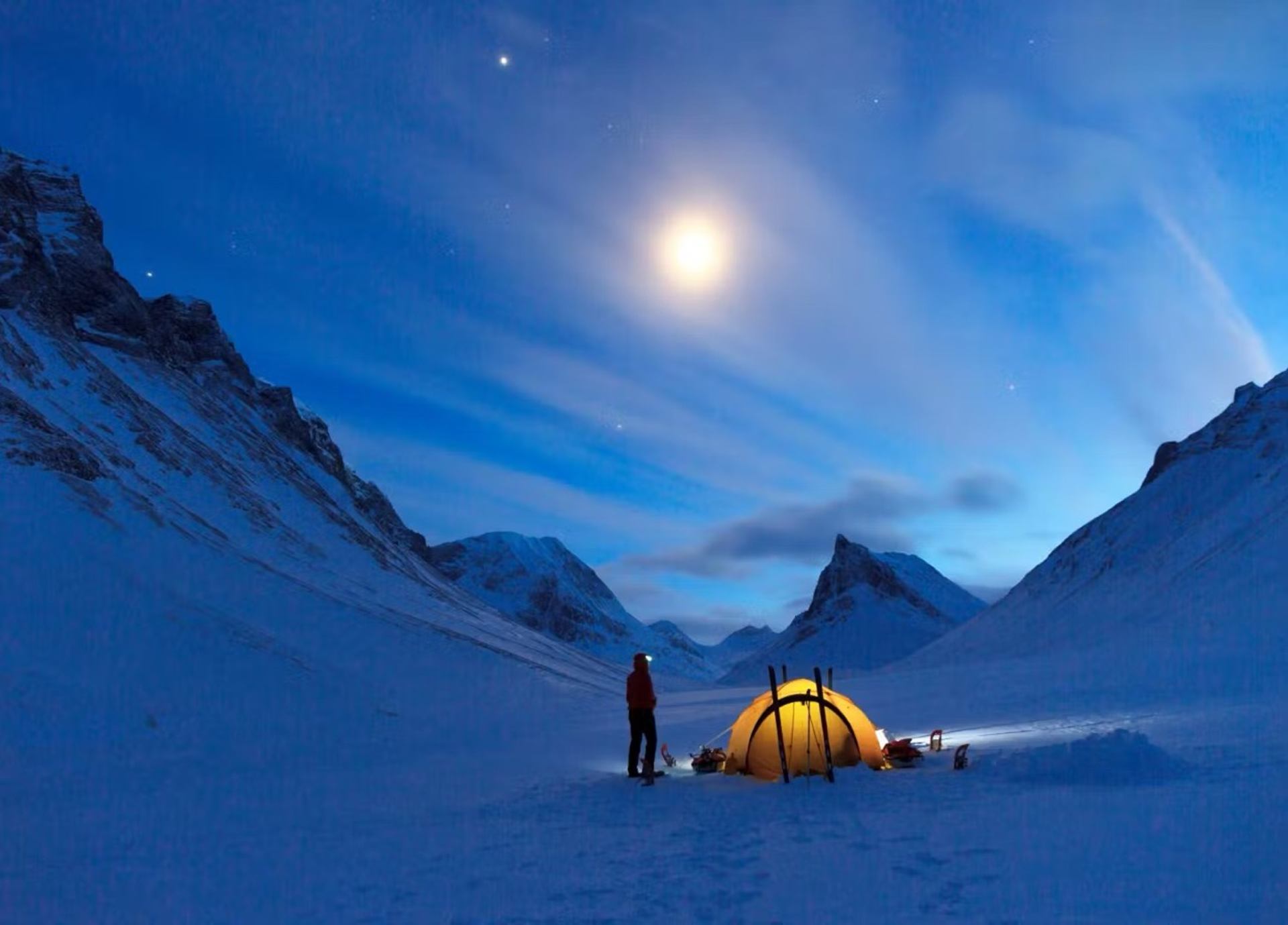Person near yellow tent in snowy mountain valley under moonlit night sky.
