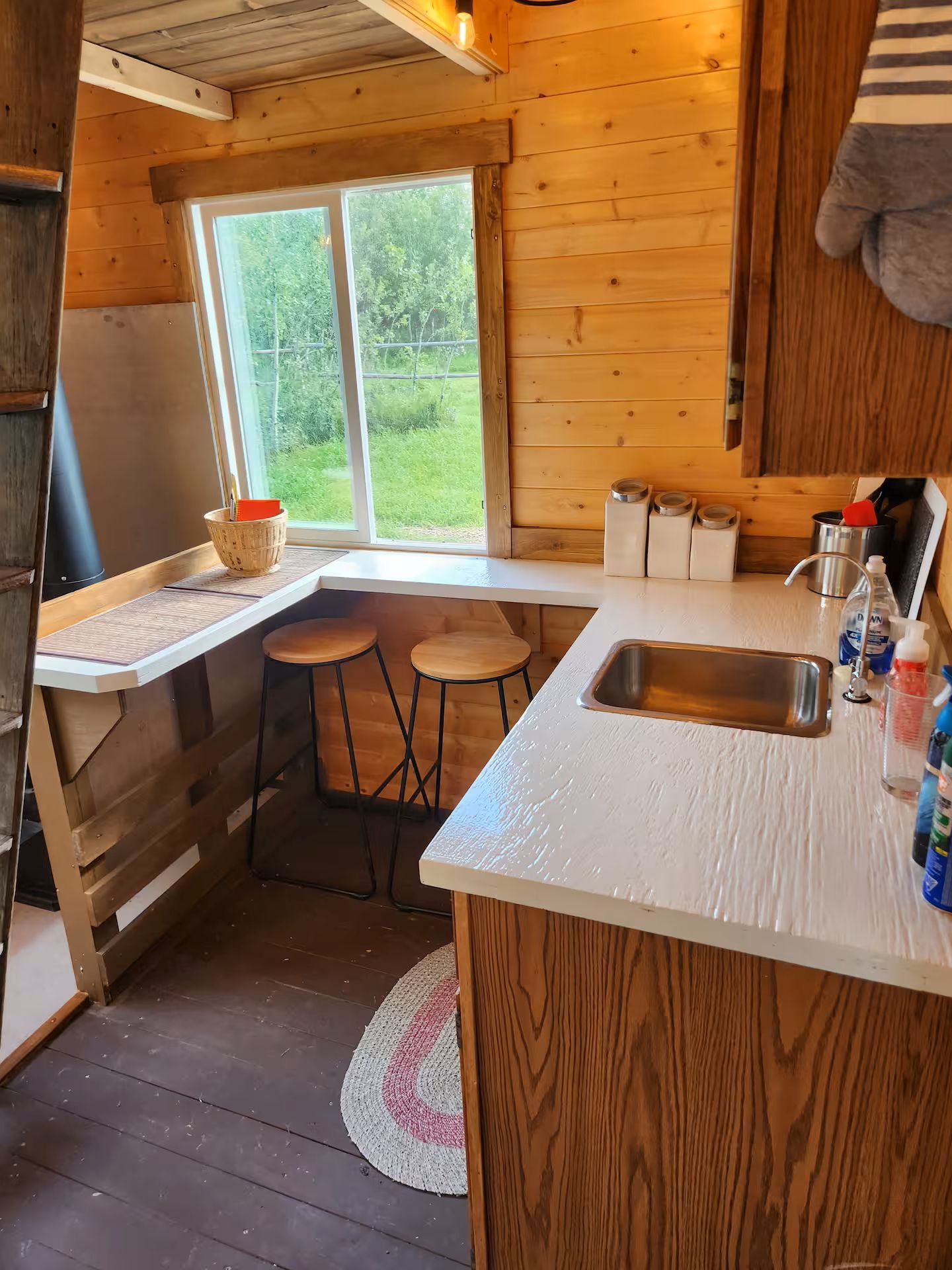 Small kitchen with wooden cabinets, white counter, and bar stools by a window.