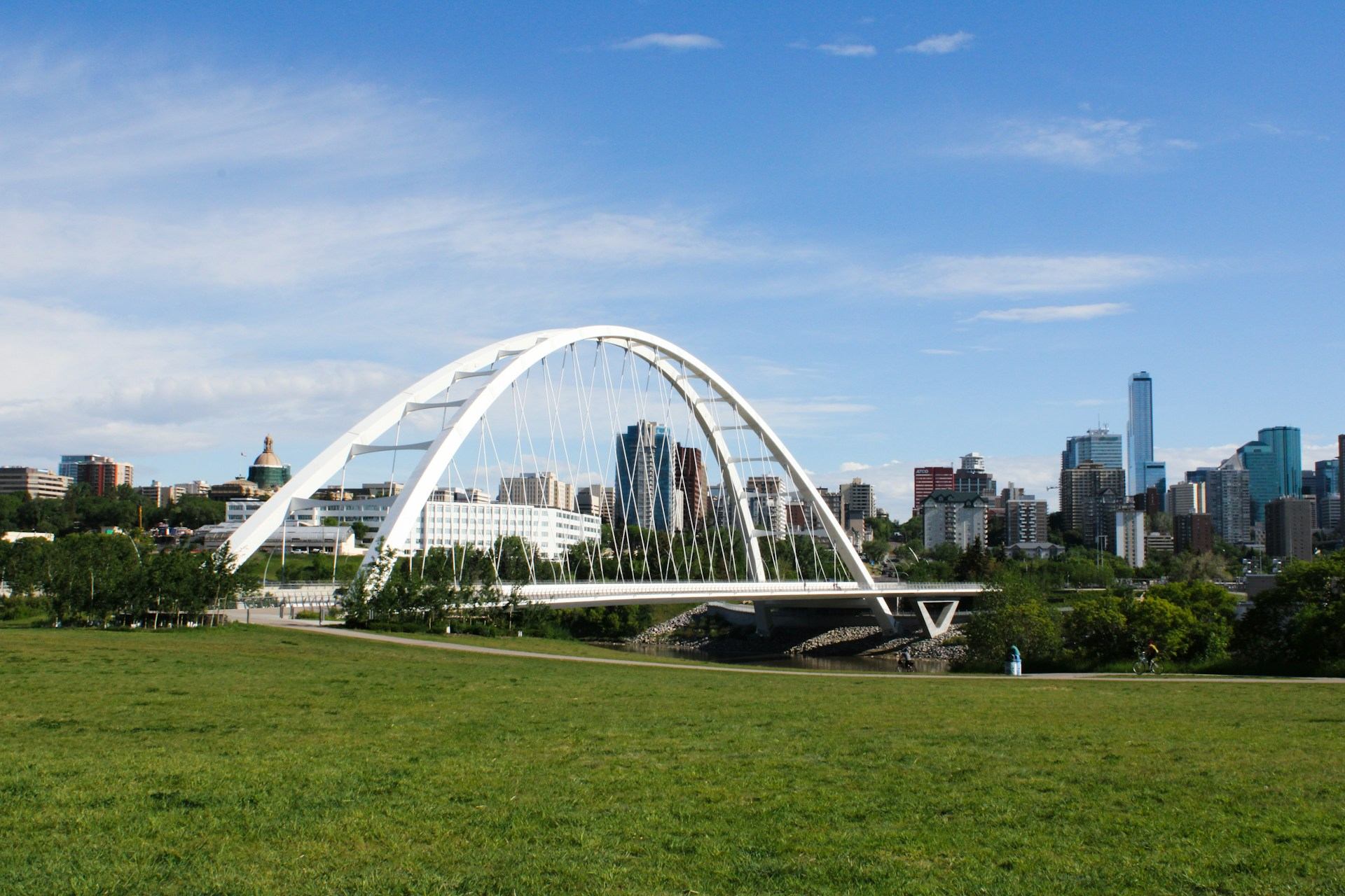 White arch bridge spans river before city skyline, seen from grassy park foreground.