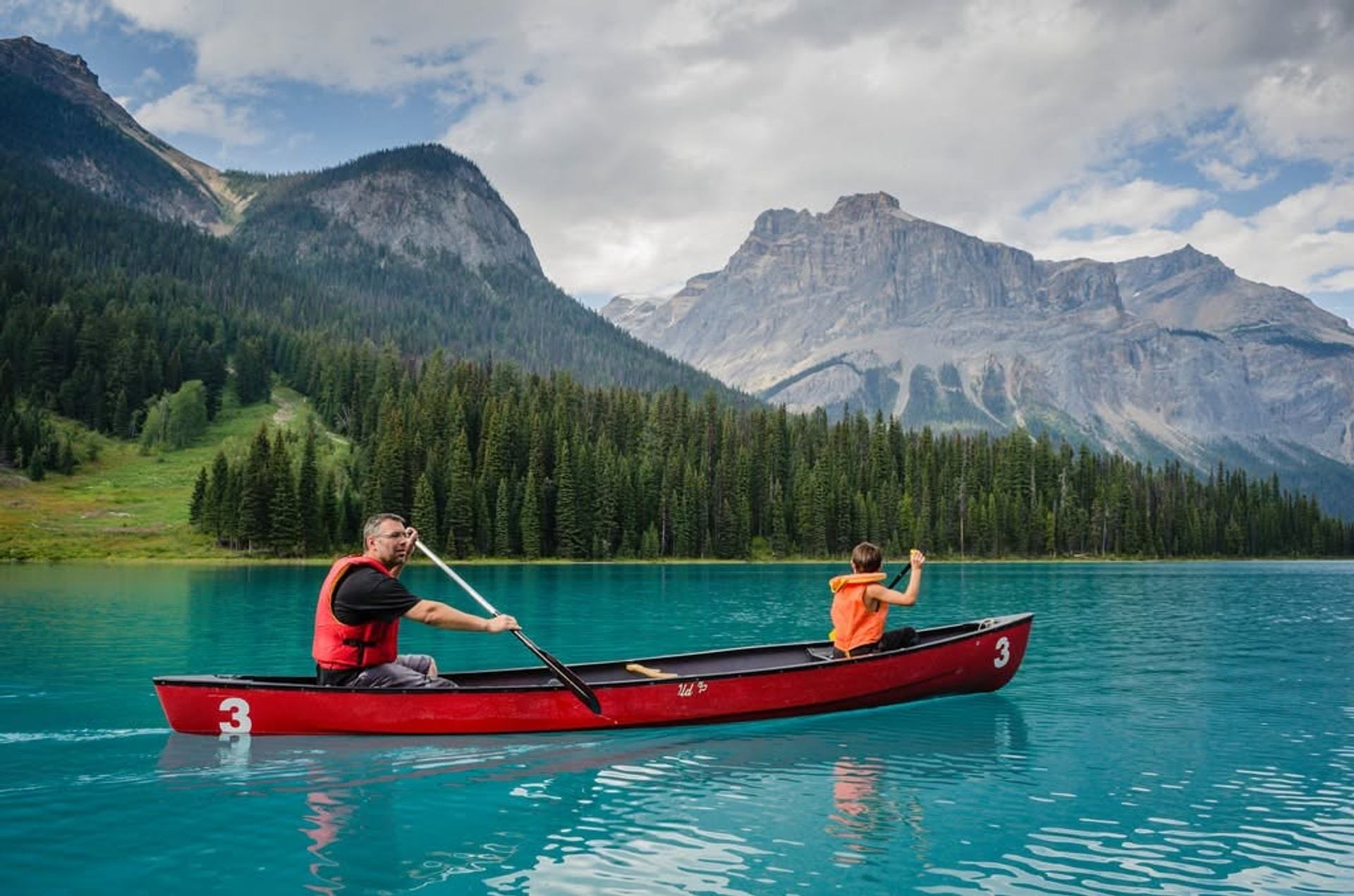 Canoeists paddle a red canoe across a turquoise mountain lake bordered by evergreens.
