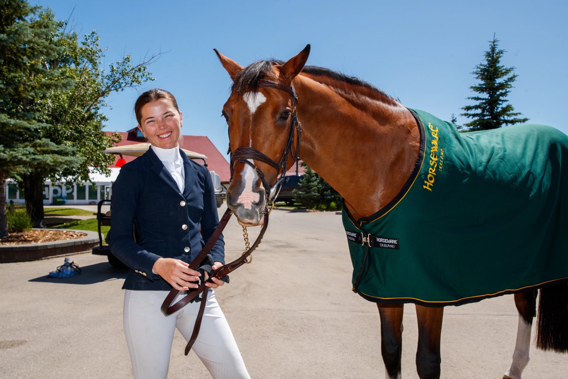 A rider stands beside a saddled horse wearing a green blanket on tournament grounds.