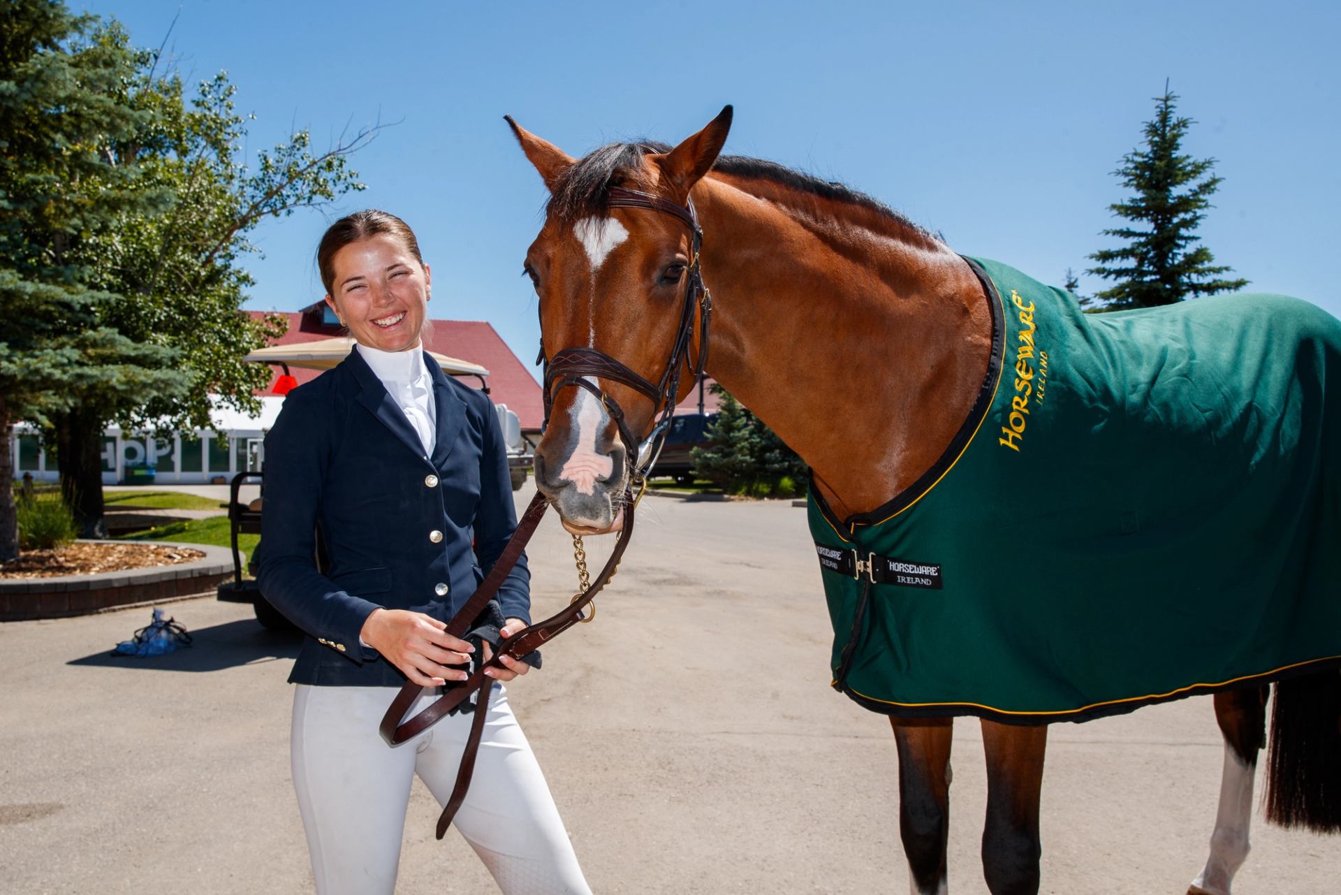 A rider stands beside a saddled horse wearing a green blanket on tournament grounds.