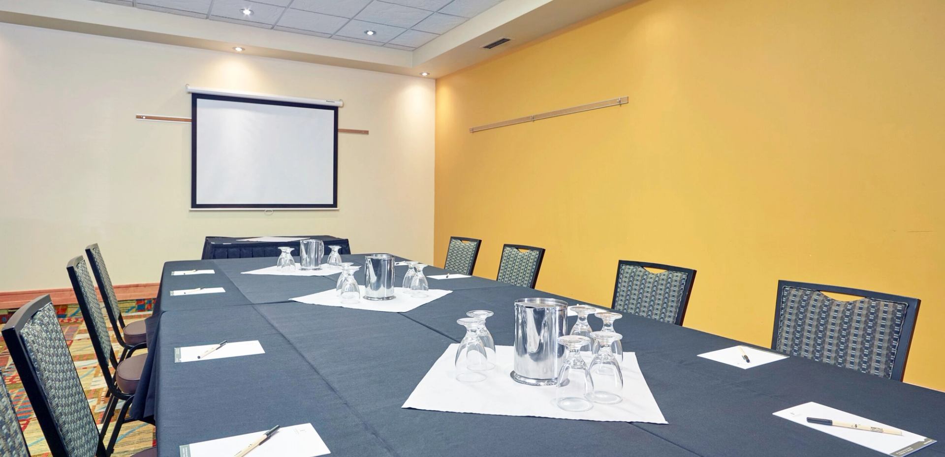 A meeting room has a long table covered with a dark tablecloth, surrounded by chairs. Glasses, notepads, and a projector screen at the front make it suitable for small meetings or presentations.