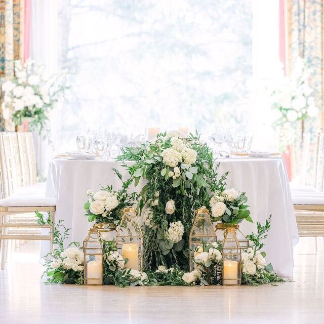 Wedding reception table with lush white florals, greenery, candles, and gold accents in a bright indoor space.