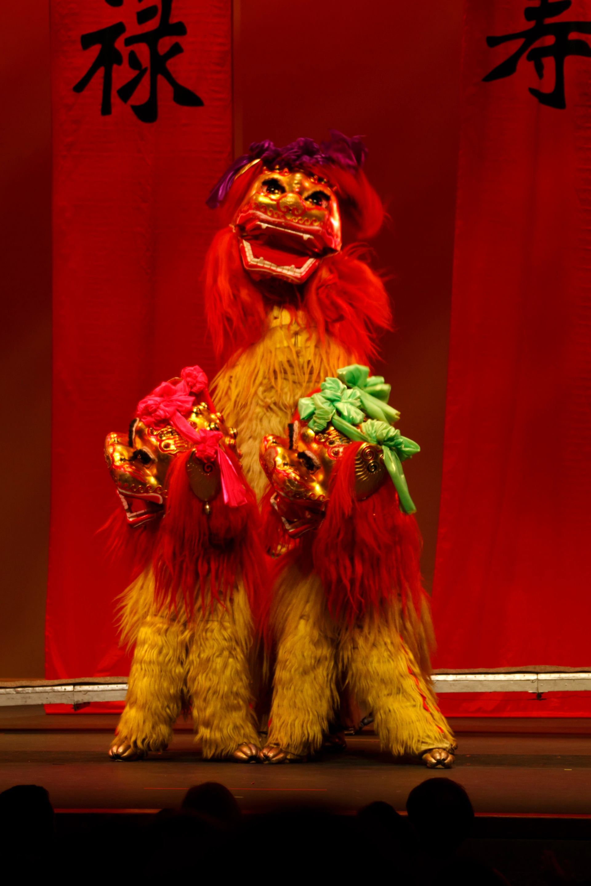 Lion dance costumes with gold masks and red fur perform on stage before red banners.