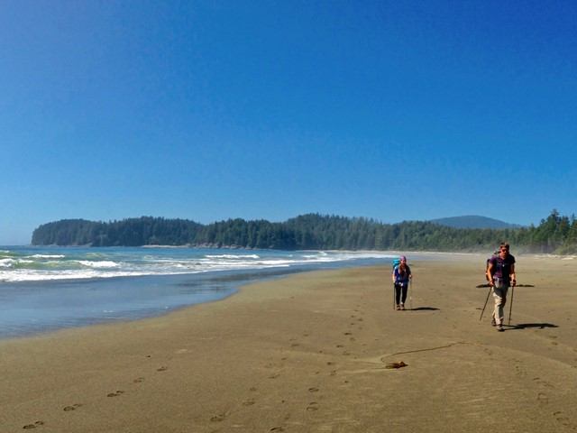 Two people hiking on a wide sandy beach beside ocean waves and forested headlands.