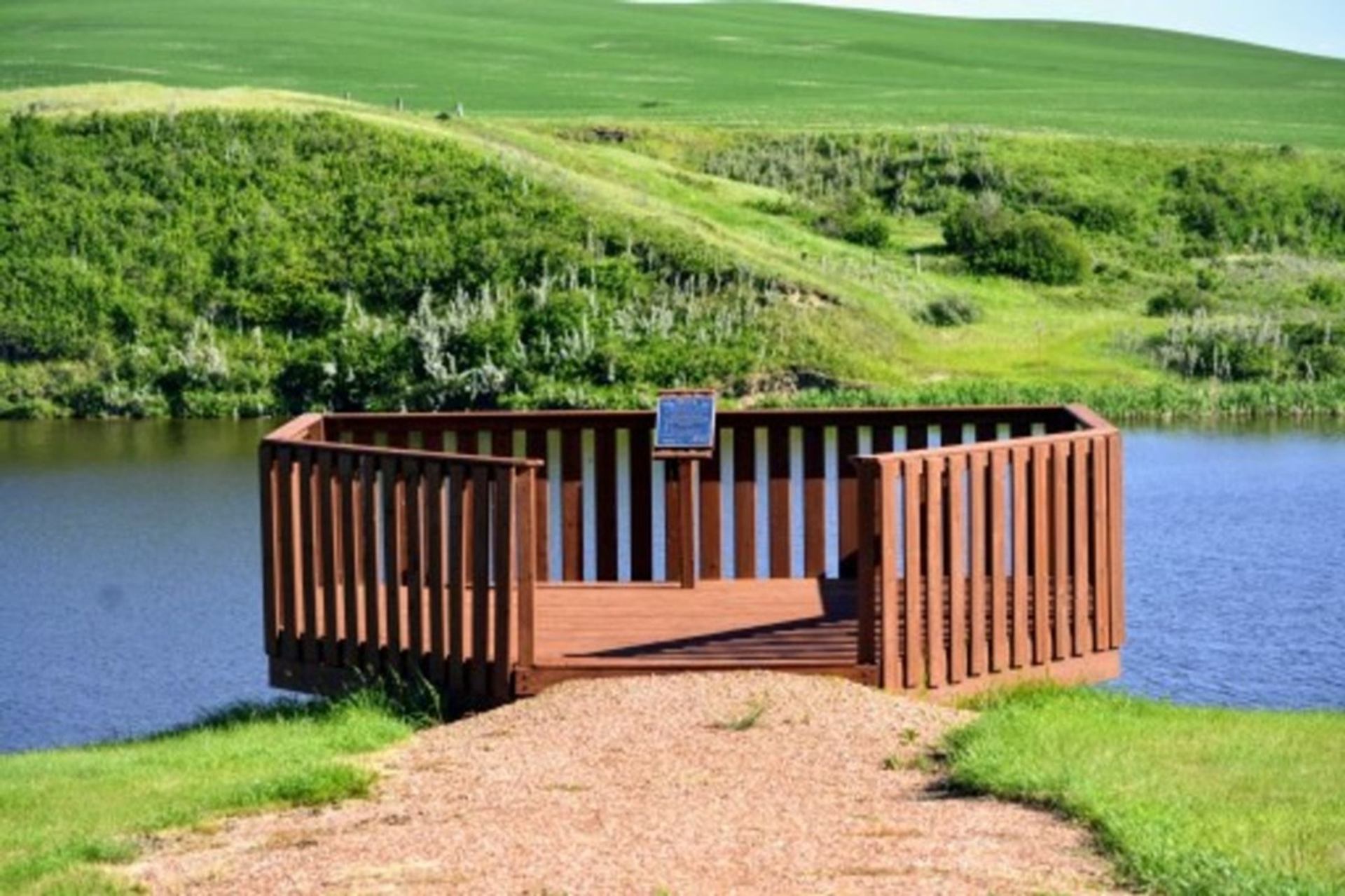 Wooden viewing platform over calm water, surrounded by green grassy hills and shrubs.