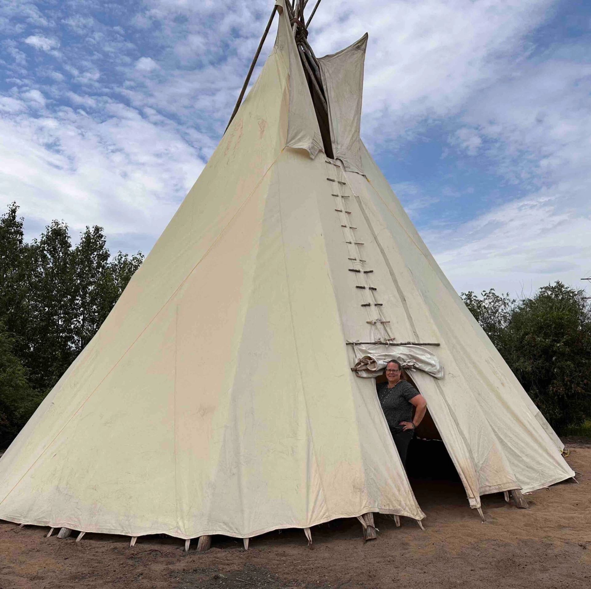Tipi tent with person standing in the open doorway under cloudy sky.