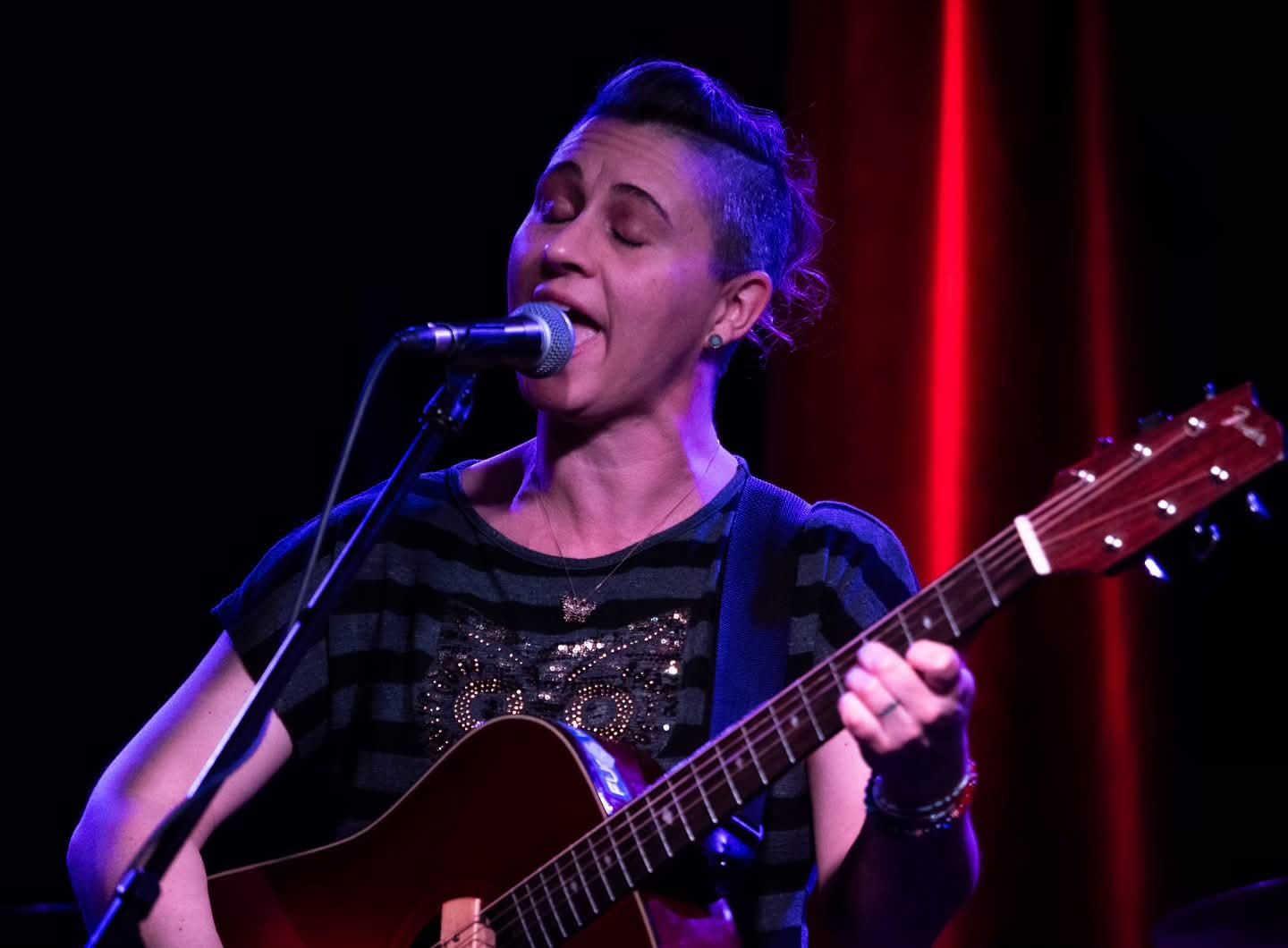 Musician sings into a microphone while playing acoustic guitar under red and purple lights.