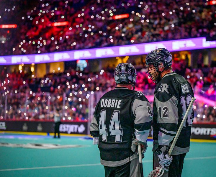 Two Calgary Roughnecks players on field, crowd lit up with cell phone lights in indoor stadium.