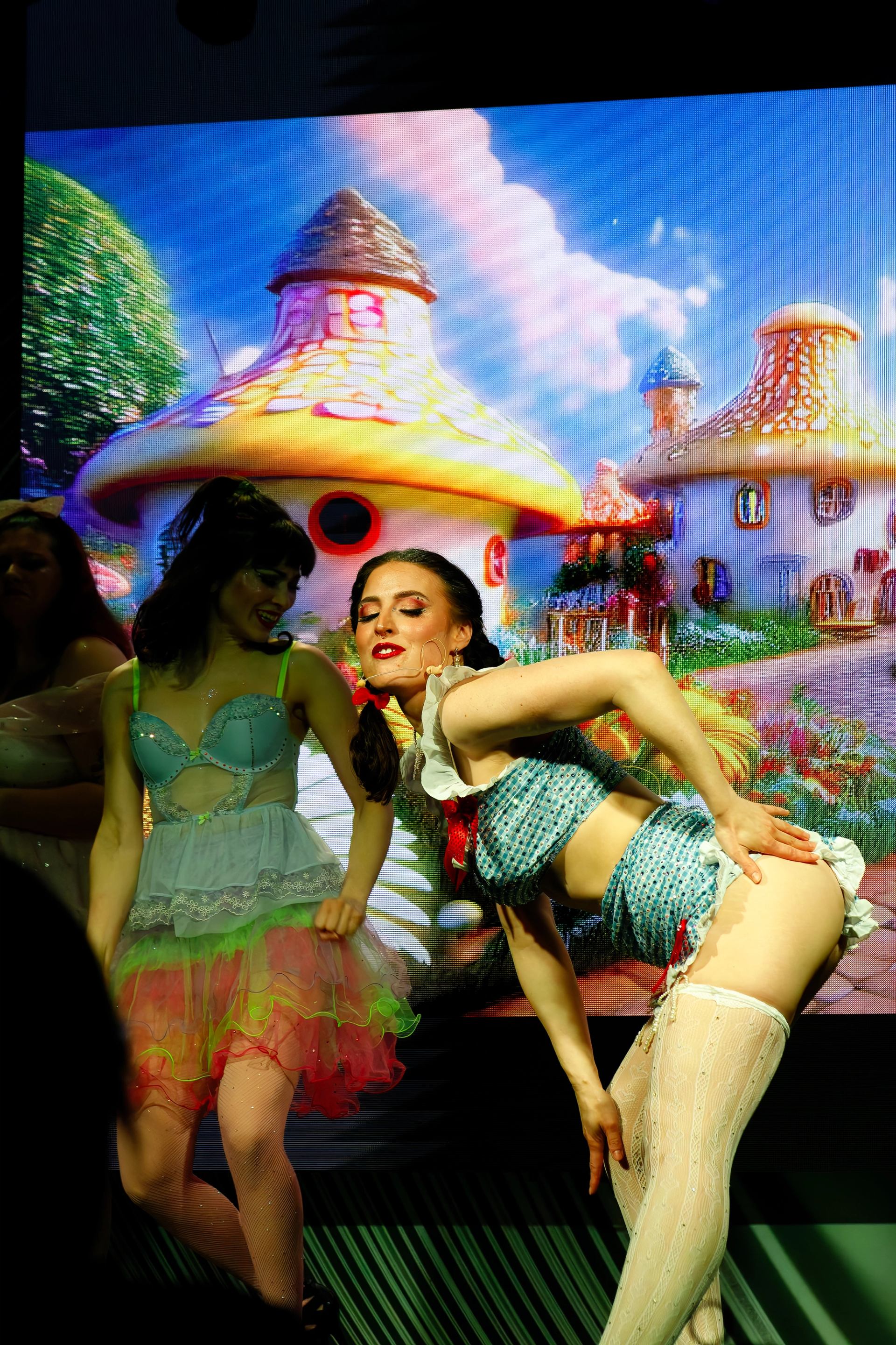 Two costumed performers on stage with fantasy mushroom village backdrop.