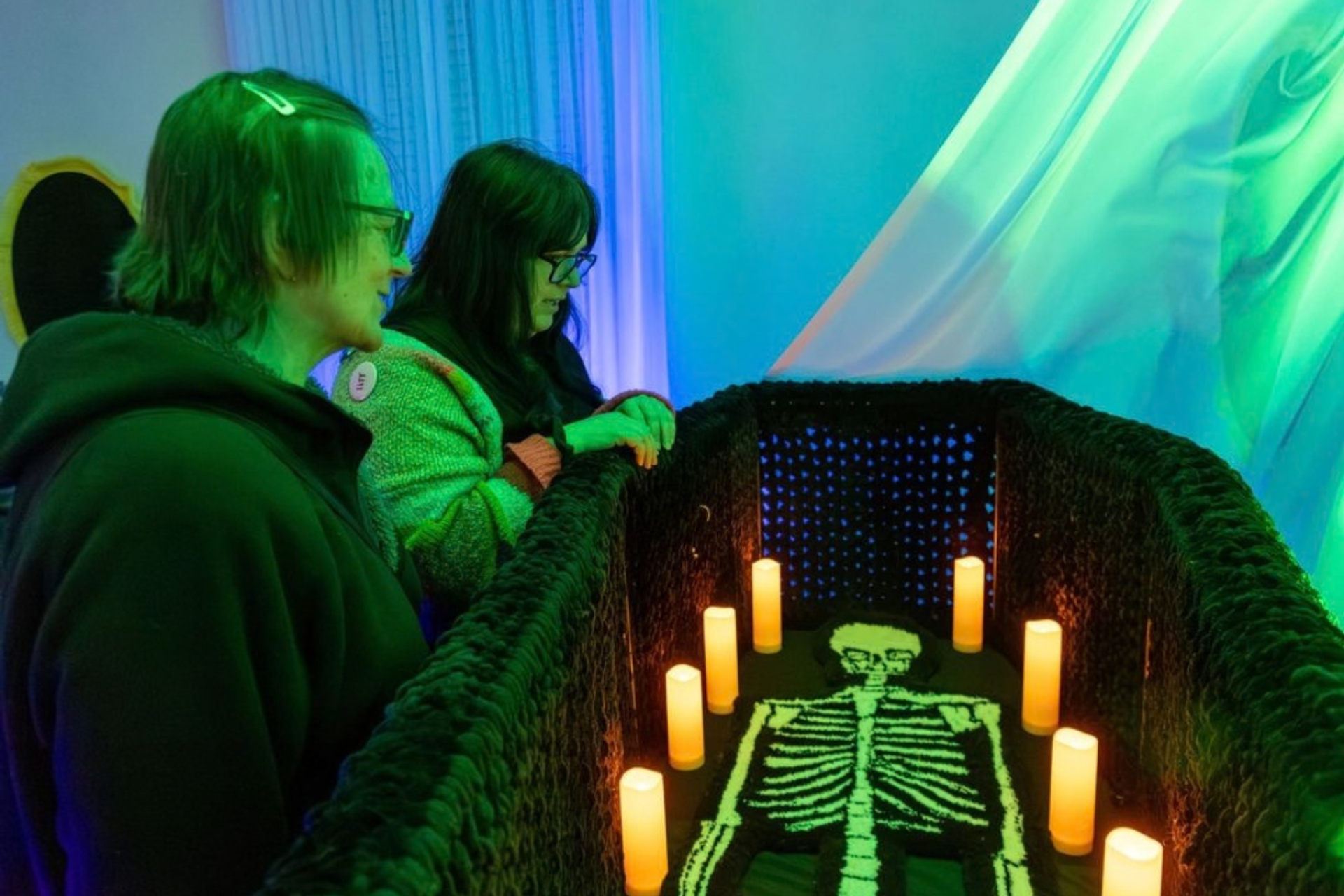 Visitors view a glowing skeleton display surrounded by candles under green and blue lights.