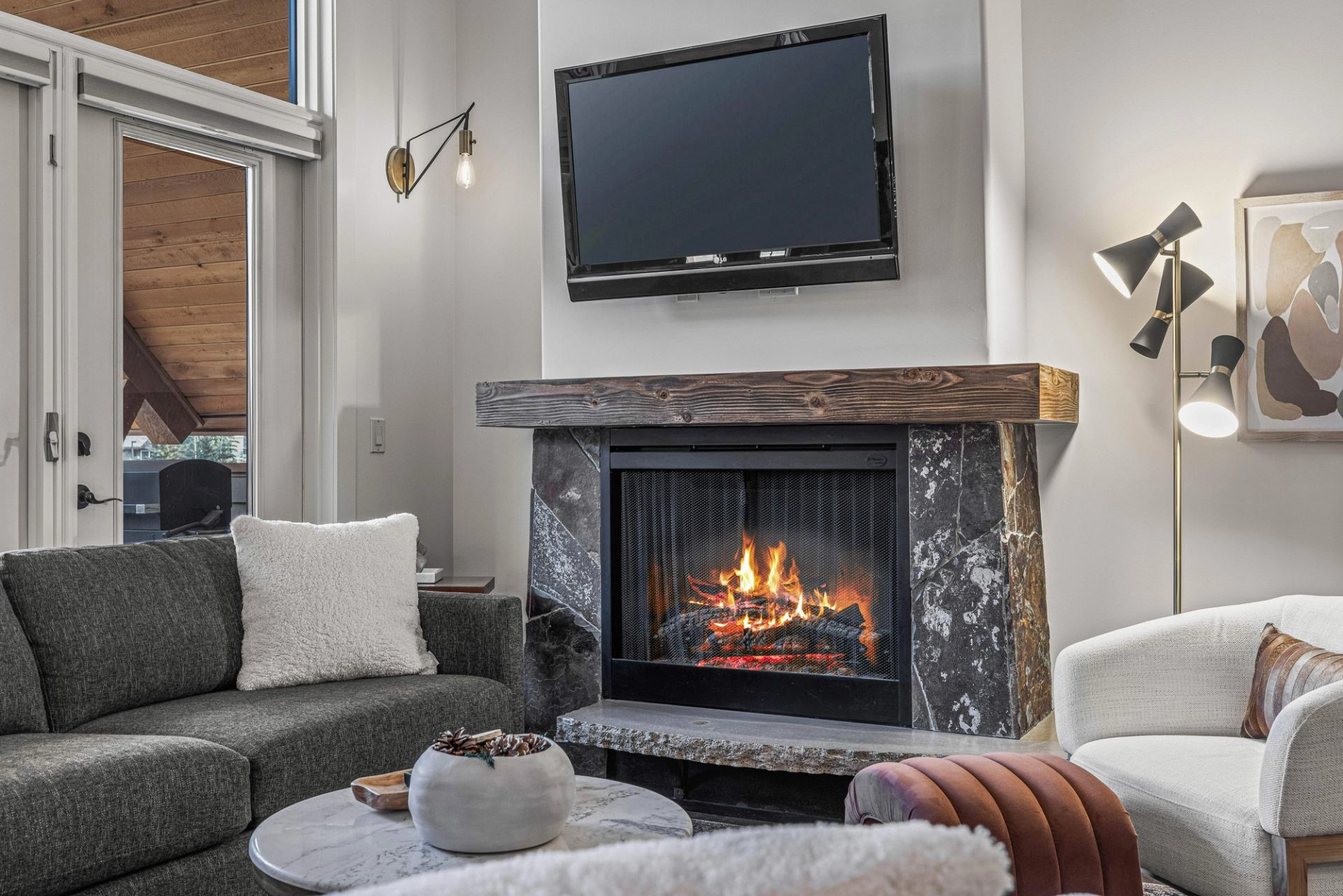 Living room with lit stone fireplace, wall-mounted TV, sofa and armchairs.