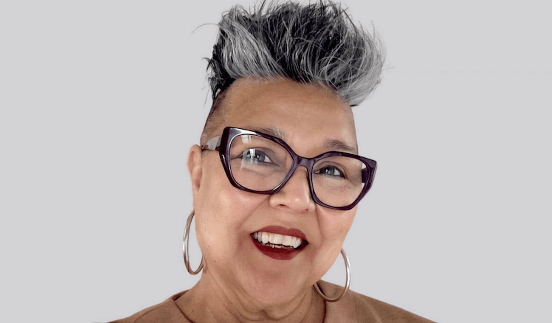 Person with short grey and black hair, large glasses and hoop earrings against grey background.