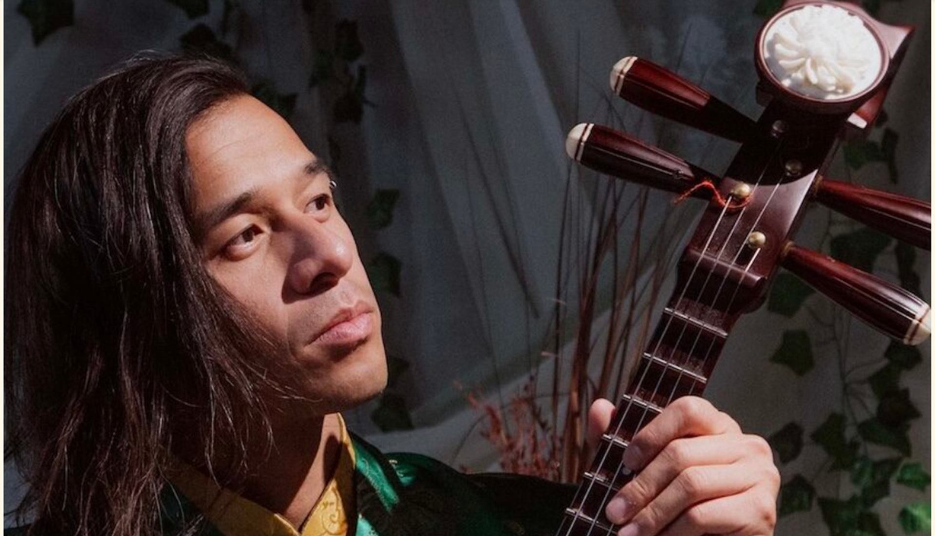 Person holding a stringed instrument with carved headstock, standing before leafy backdrop.