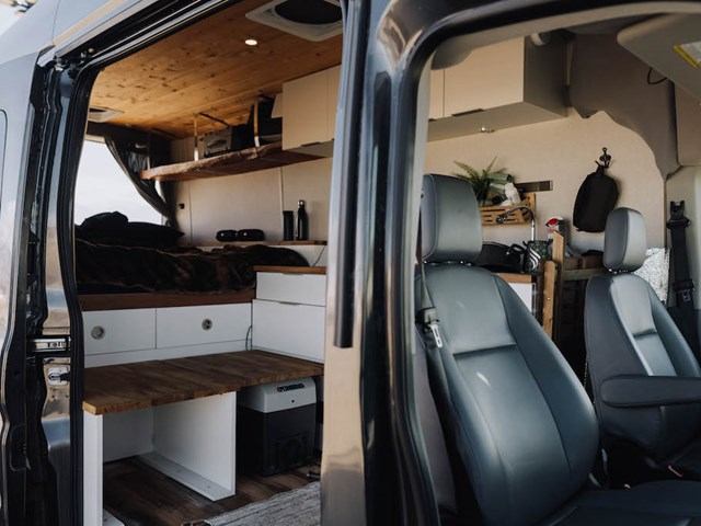 Campervan interior with wood ceiling, bed, shelves, and front seats.