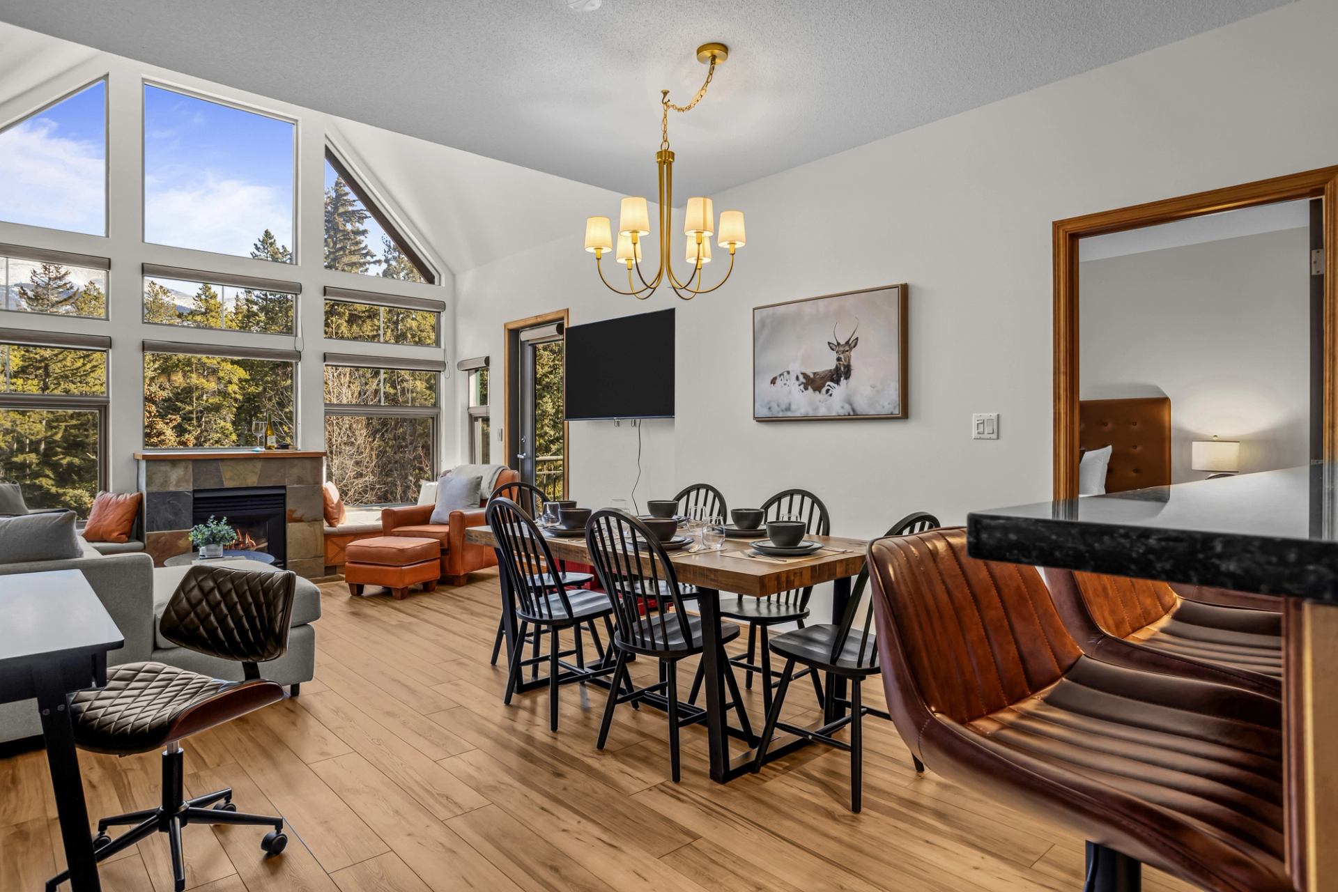 An open living and dining area includes a dining table with seating, leather lounge chairs, a fireplace, large windows, and a wall-mounted television with forest views outside.