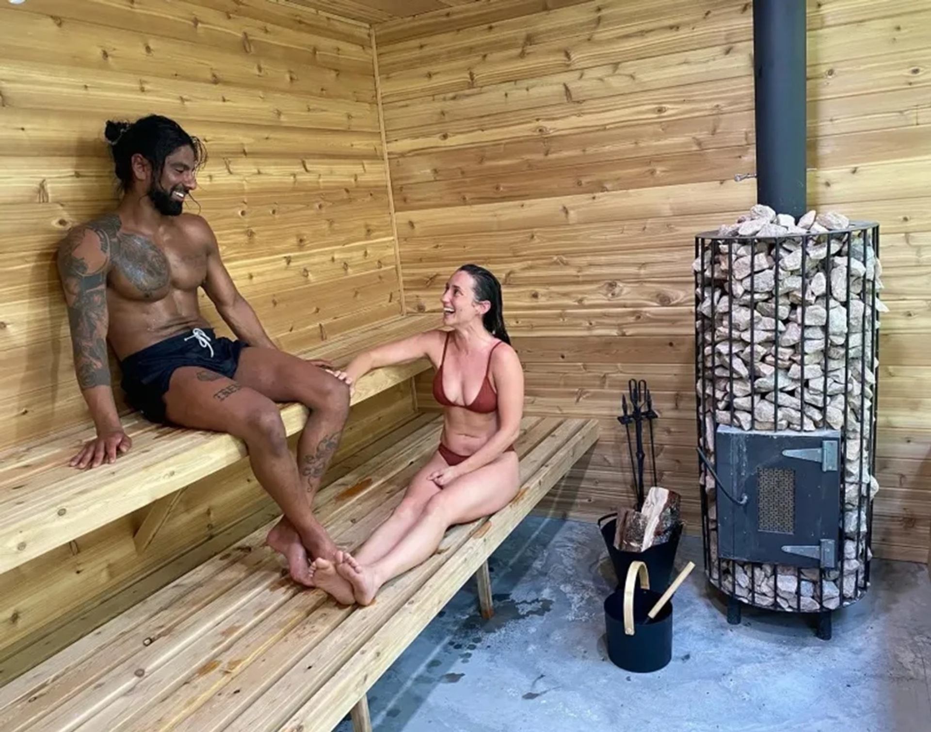 Two people sit on wooden sauna benches beside a stone-filled wood stove.