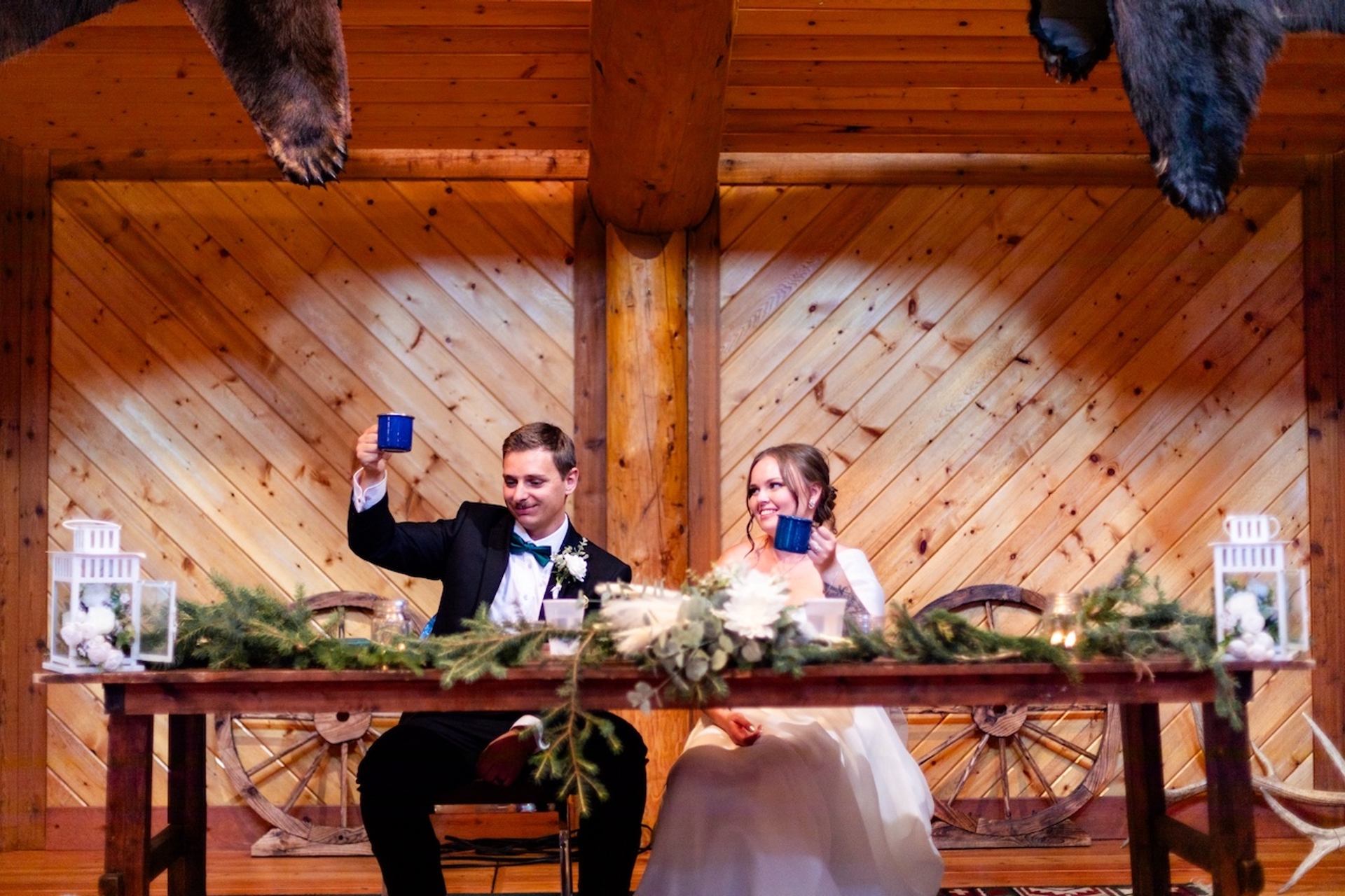 Two people seated at a decorated wooden table inside a rustic reception hall.