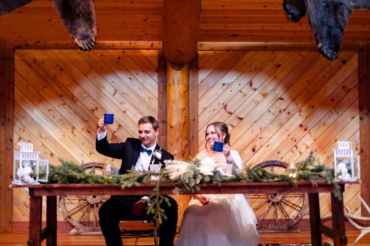 Two people seated at a decorated wooden table inside a rustic reception hall.