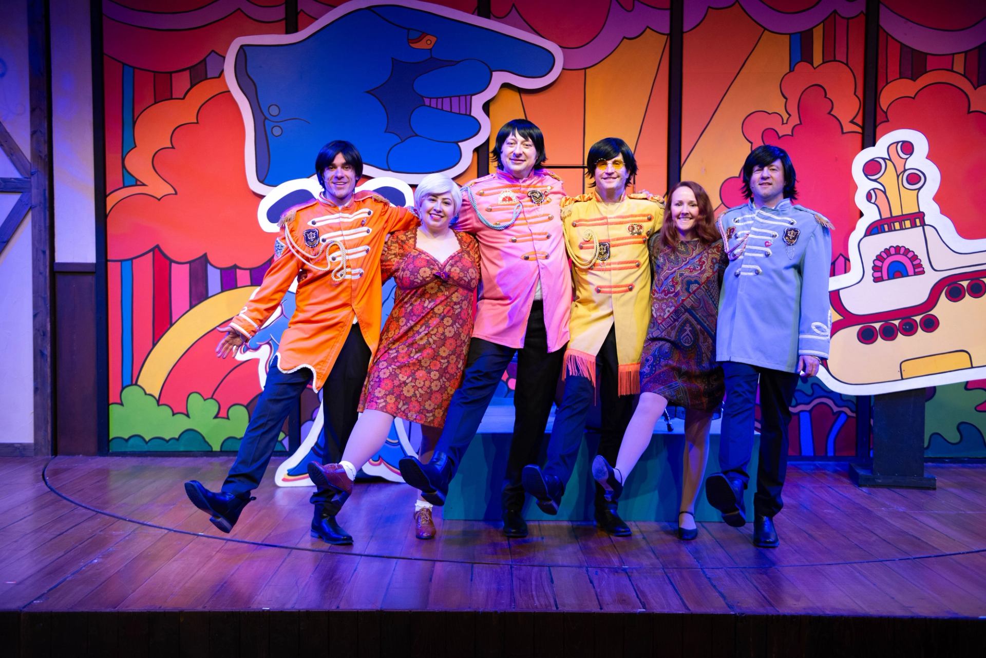 Performers in colourful Sgt. Pepper costumes kick in unison on a psychedelic stage backdrop.