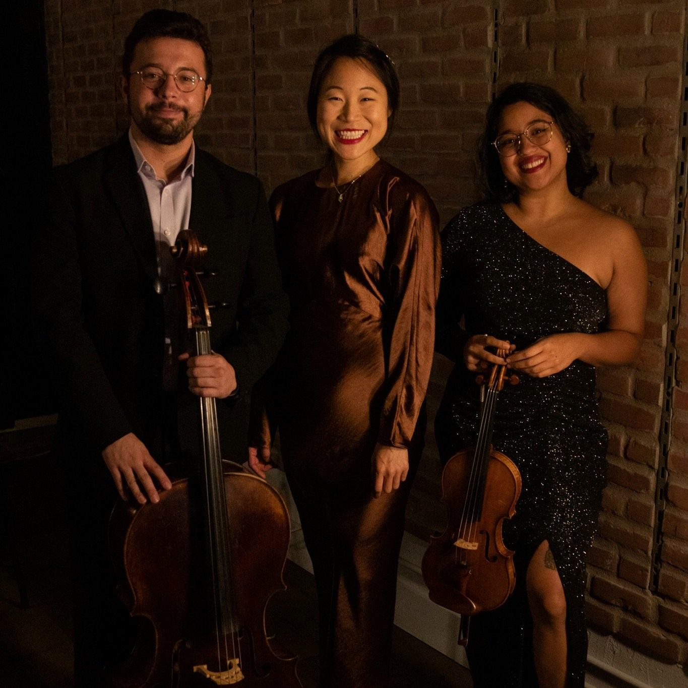 Three formally dressed musicians stand indoors, holding a cello and a violin against brick walls.