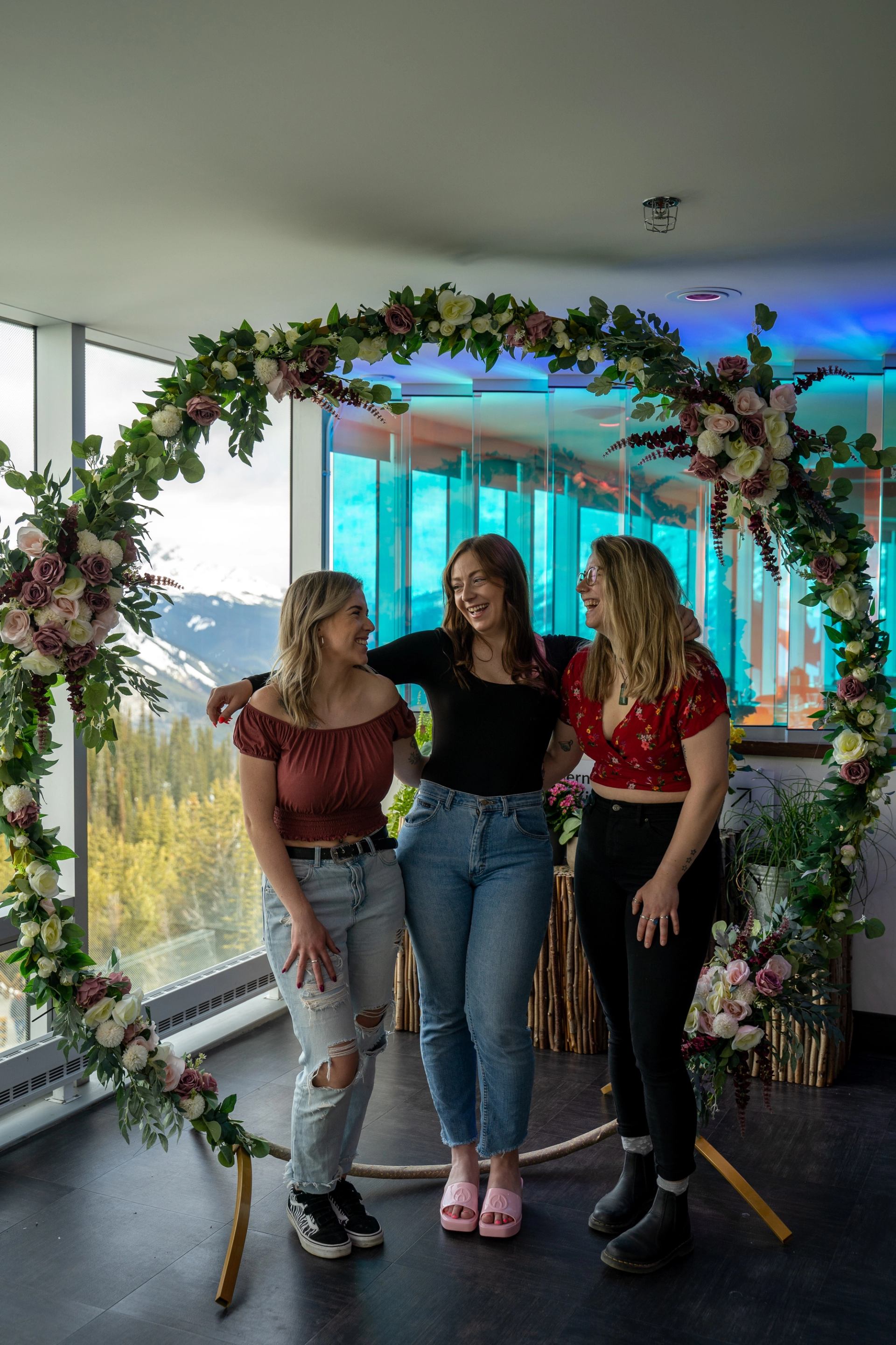Three people standing together under a large floral arch with mountains visible through the windows.