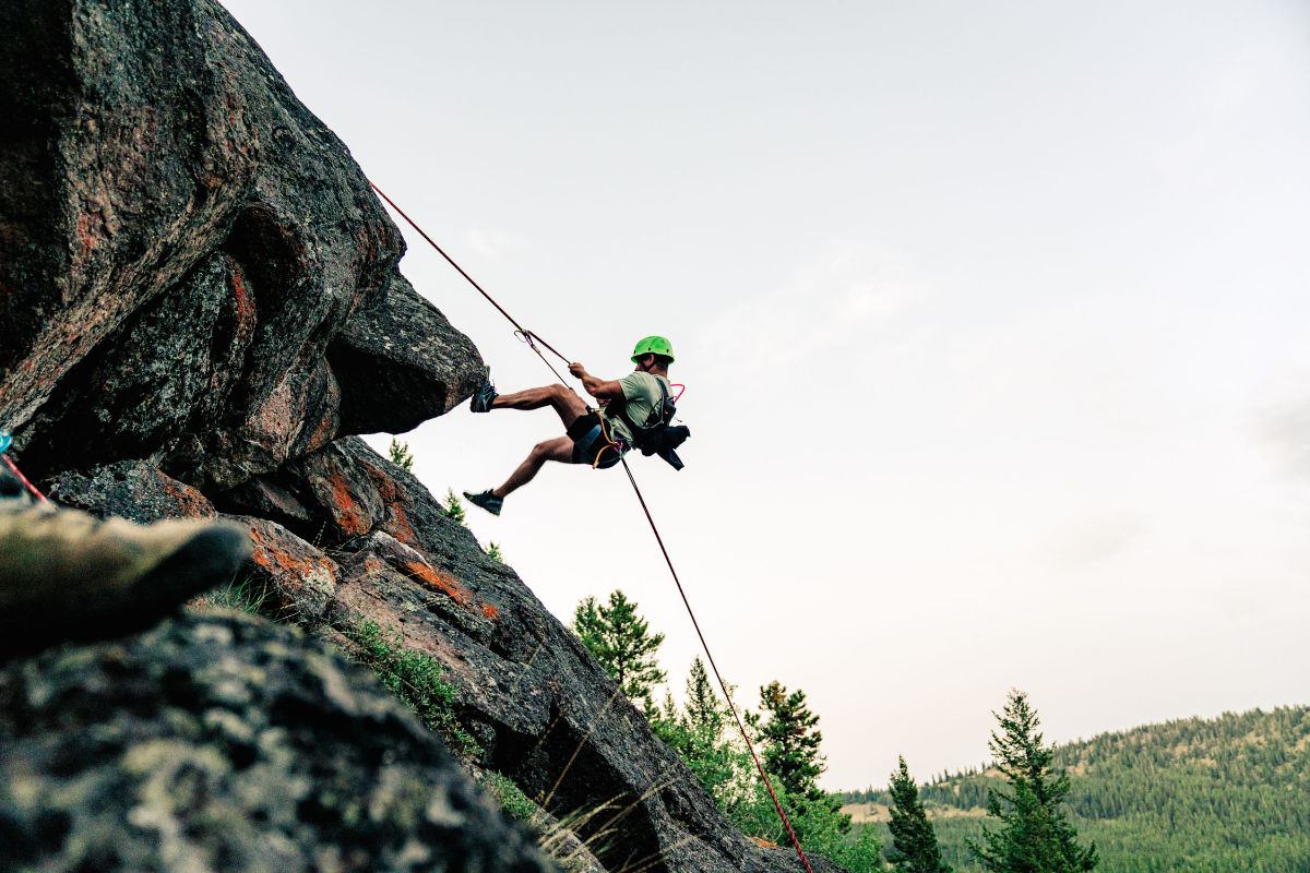 Rock climber in helmet rappels down steep cliff above forested valley landscape.