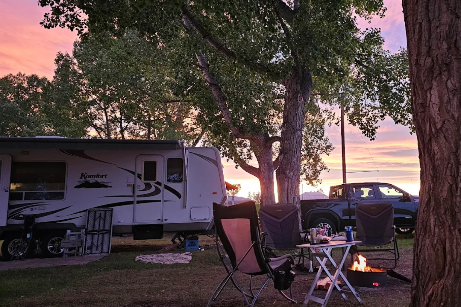 Diamond Valley Municipal Campground showcases a peaceful evening campsite with a travel trailer and pickup truck parked under large trees, glowing in a colorful sunset sky. In the foreground, the campground highlights cozy seating and a lit fire pit with a small table set up for relaxing outdoors.