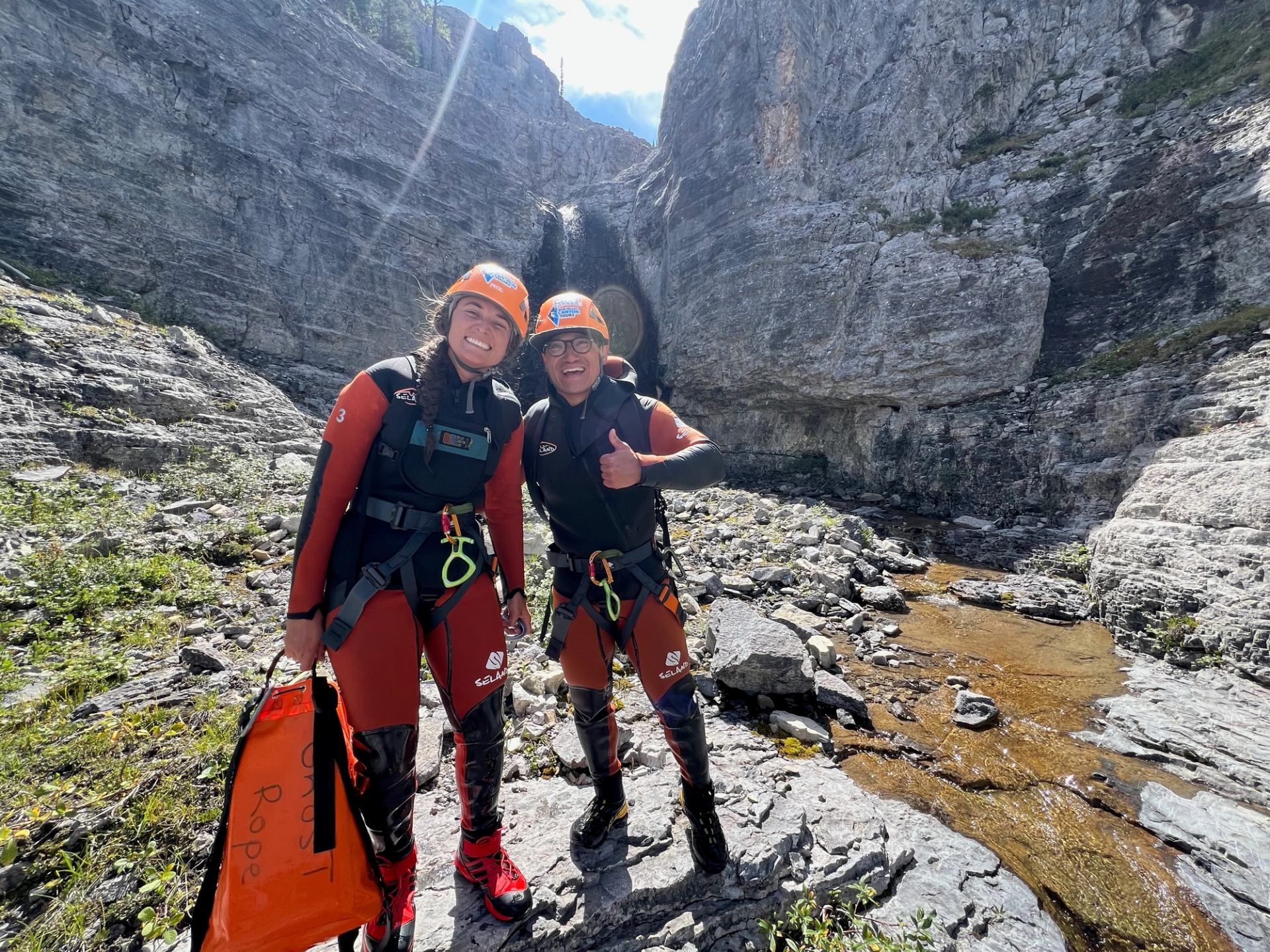 Two canyoning adventurers in helmets and gear standing on rocky terrain near a stream.