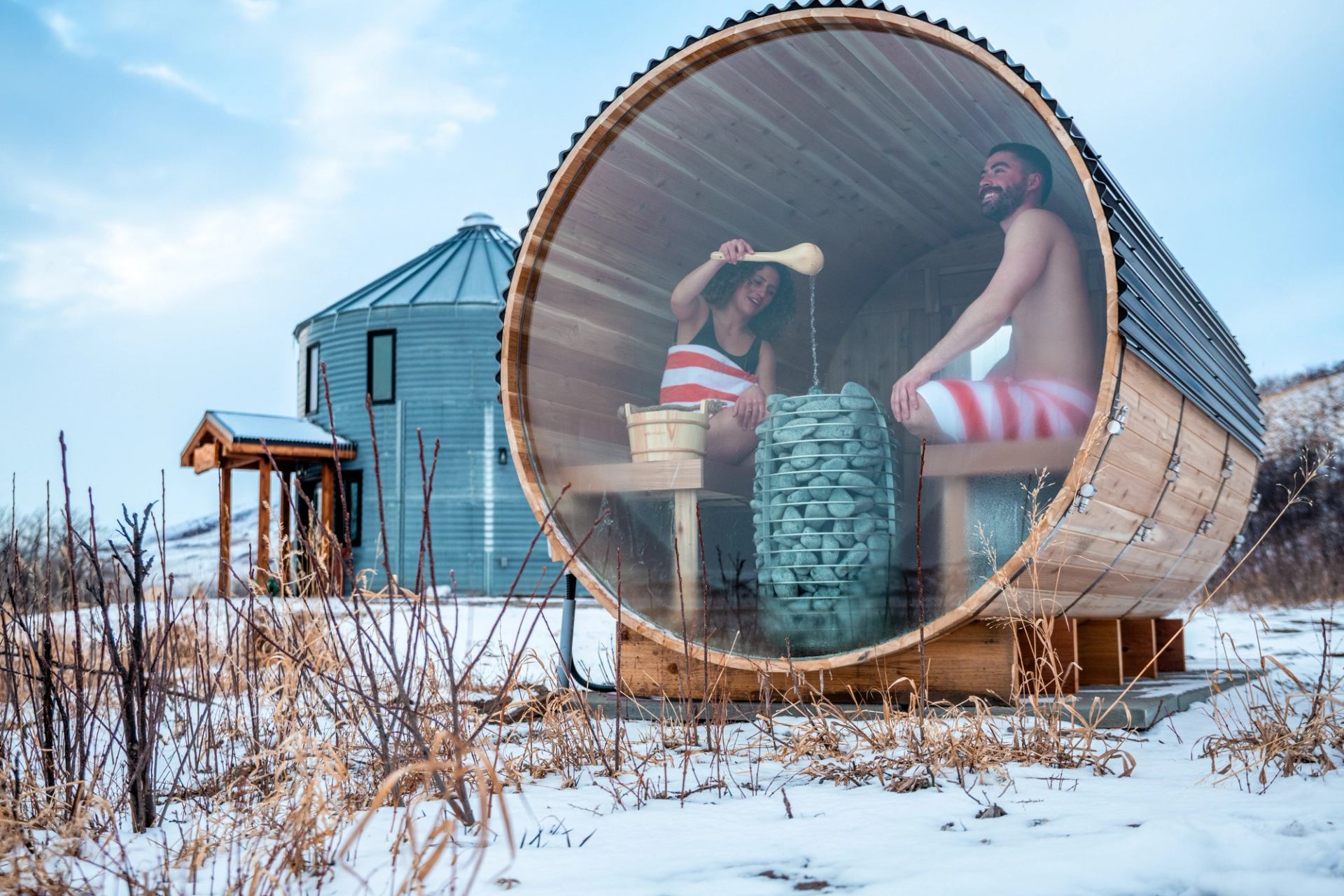 People in striped towels pour water over stones inside a barrel sauna.