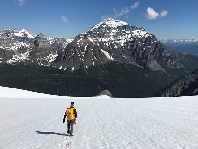 Mountaineer in orange jacket ascending a snowy slope with alpine peaks in the background.