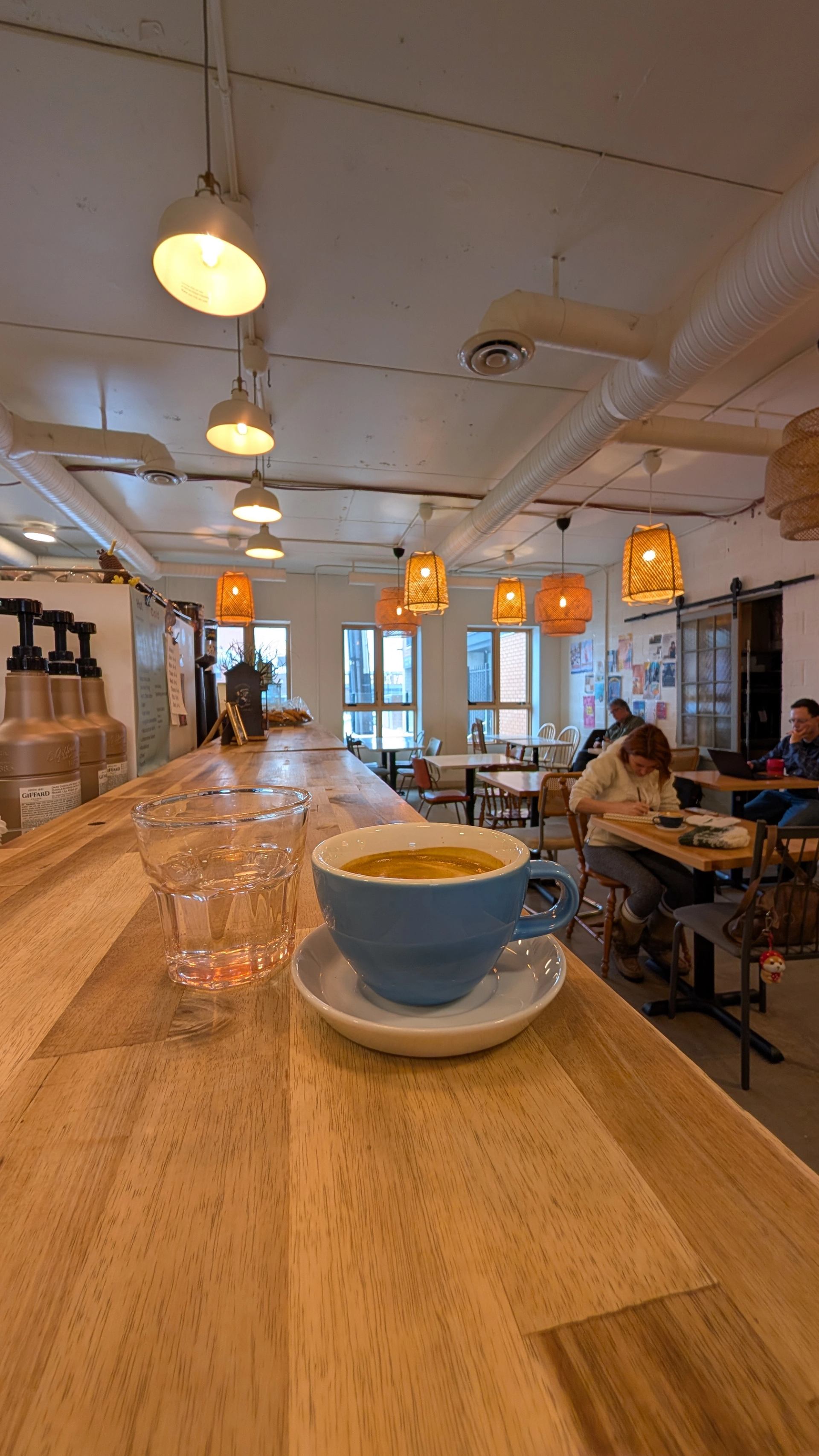 Blue coffee cup and water glass on wooden counter in busy café interior.