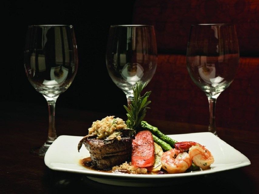A plate of steak with a topping, roasted shrimp, asparagus, and red pepper, garnished with rosemary, in front of three empty wine glasses.
