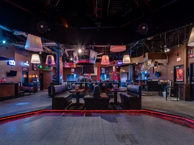 A spacious nightclub interior features booth seating, a central dance area outlined with red lighting, and hanging pendant lights. The room is dimly lit with colorful accents and a bar visible in the background.