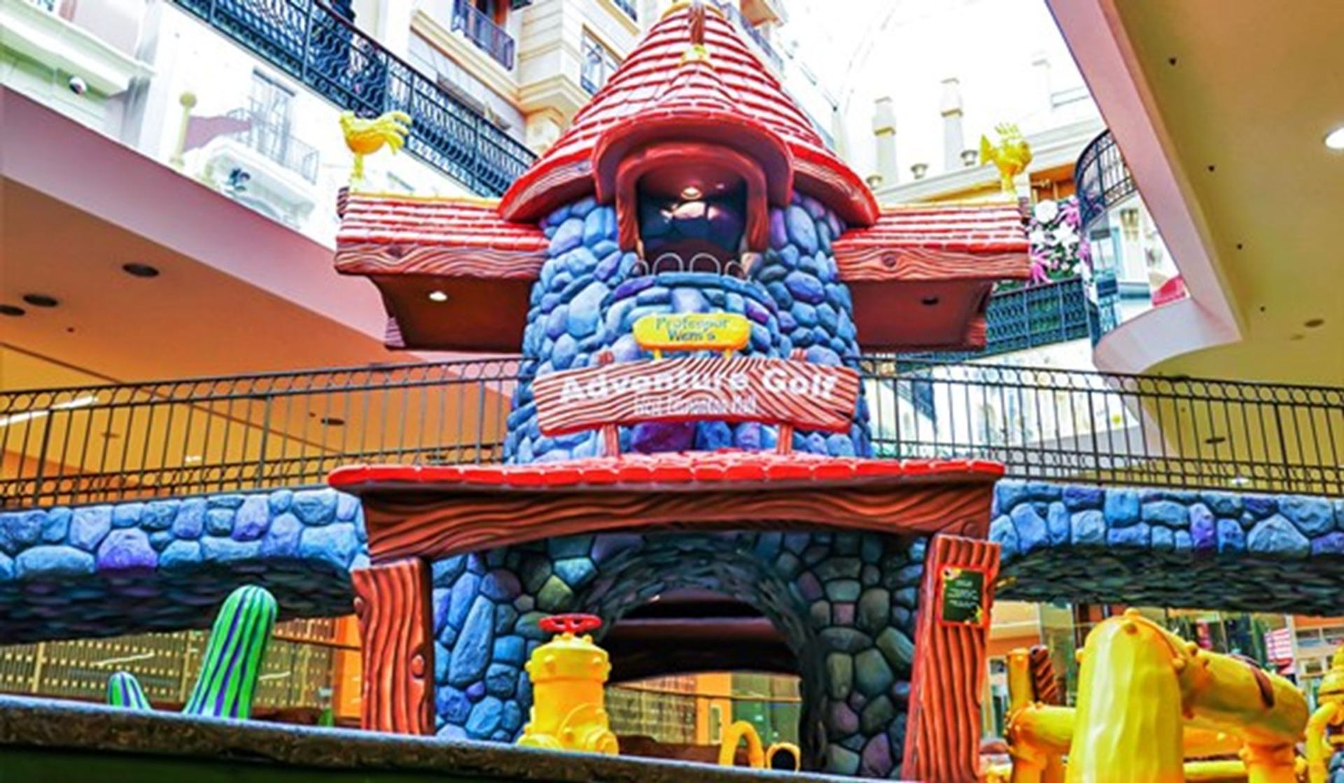 Indoor mini-golf course with colourful cartoon-style stone tower and wooden bridge structure.