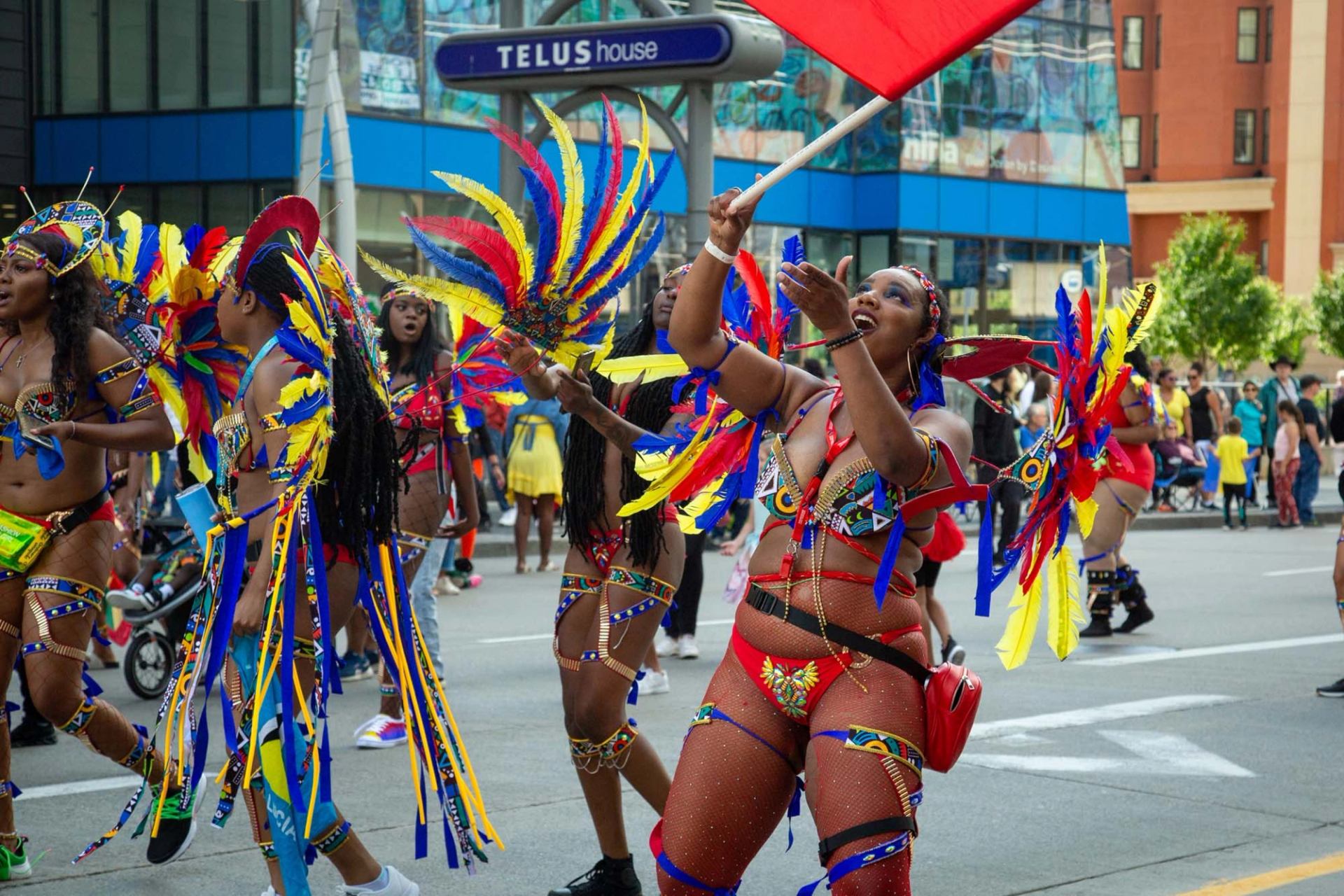 Parade performers in colourful feathered costumes dance on city street near TELUS House sign.