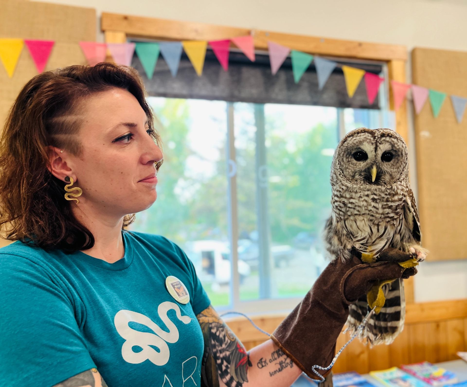 Person in teal shirt holding barred owl on gloved hand indoors.