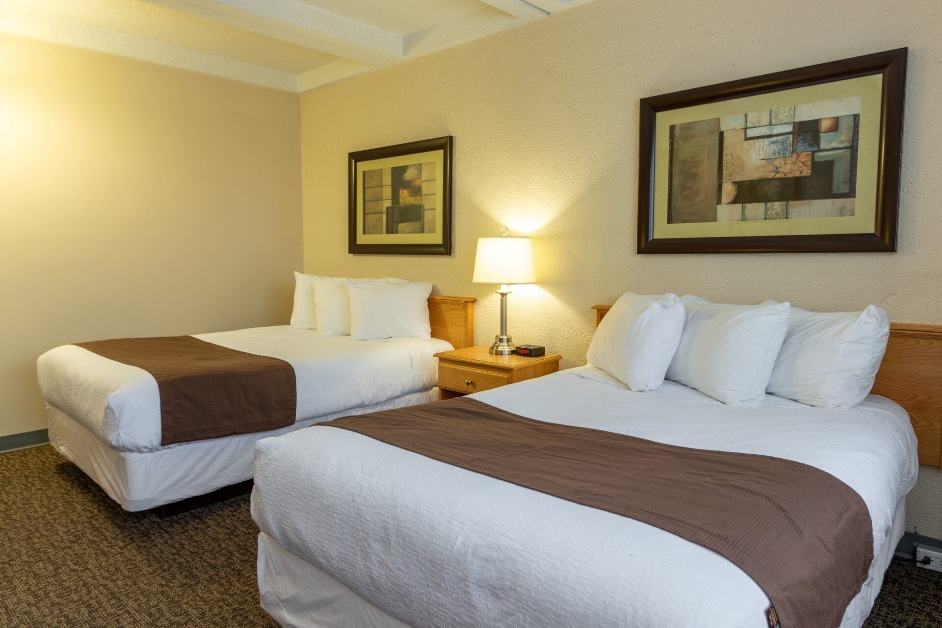Hotel room with two beds, nightstand, lamp and framed artwork on wall.