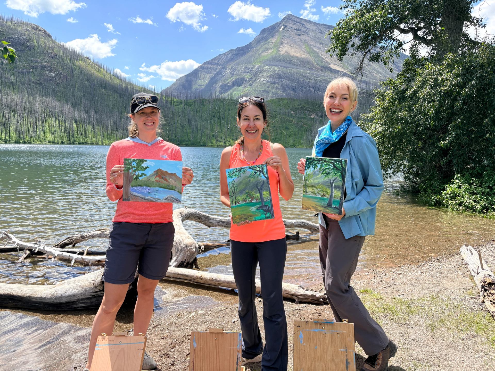 Three people hold landscape paintings beside a mountain lake with trees and driftwood.