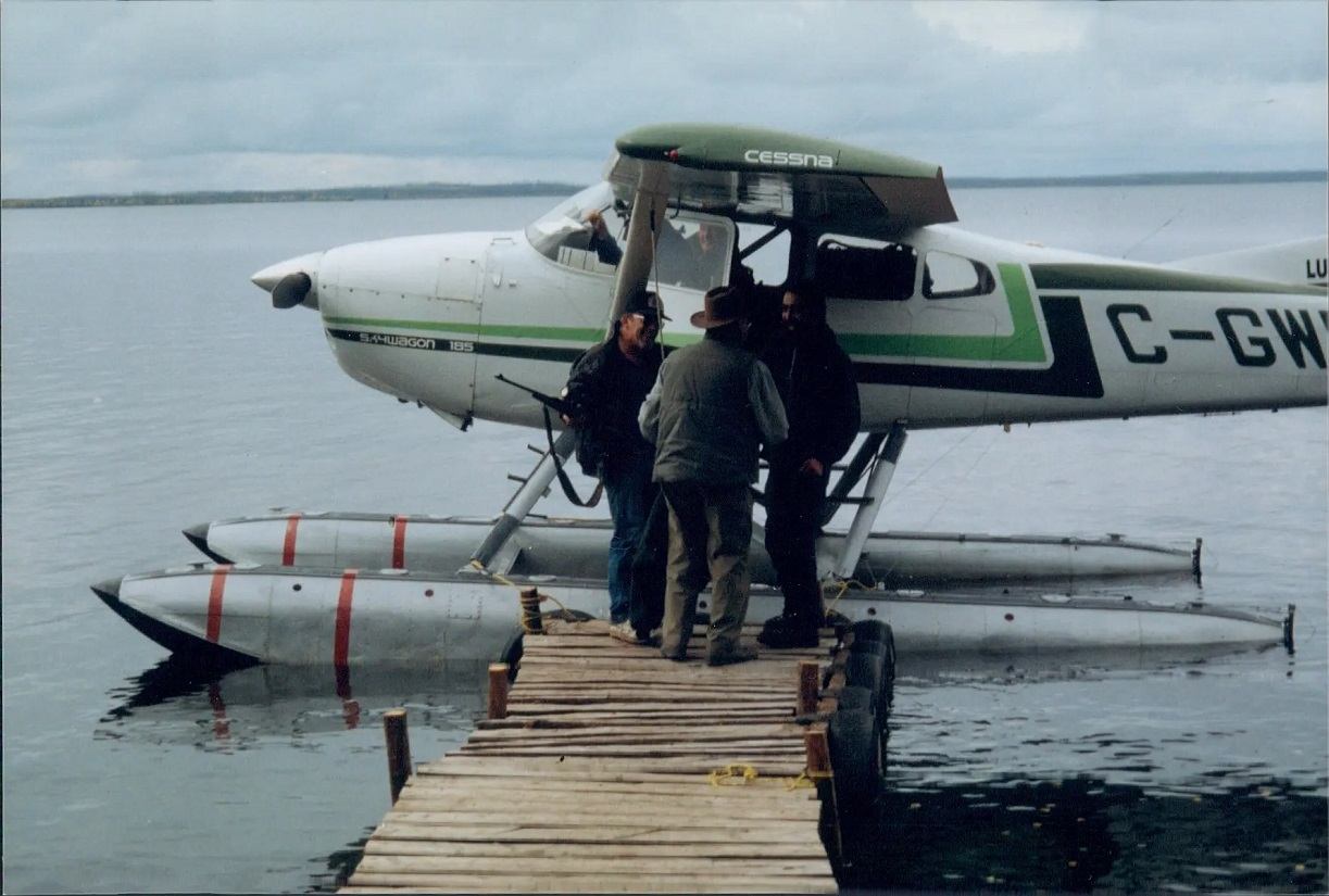 Floatplane docked at a wooden pier on a calm lake with three people standing near the aircraft.