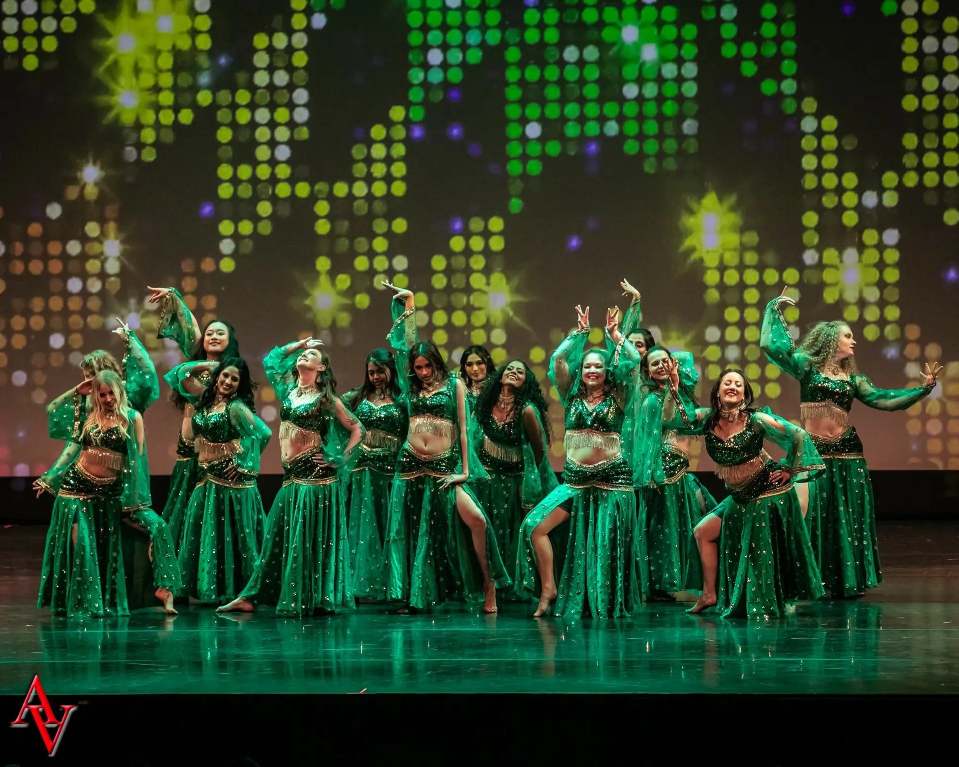 A group of dancers in matching green, glittery costumes posing together on stage in front of a colorful, abstract light-pattern backdrop, suggesting the end of a choreographed performance.
