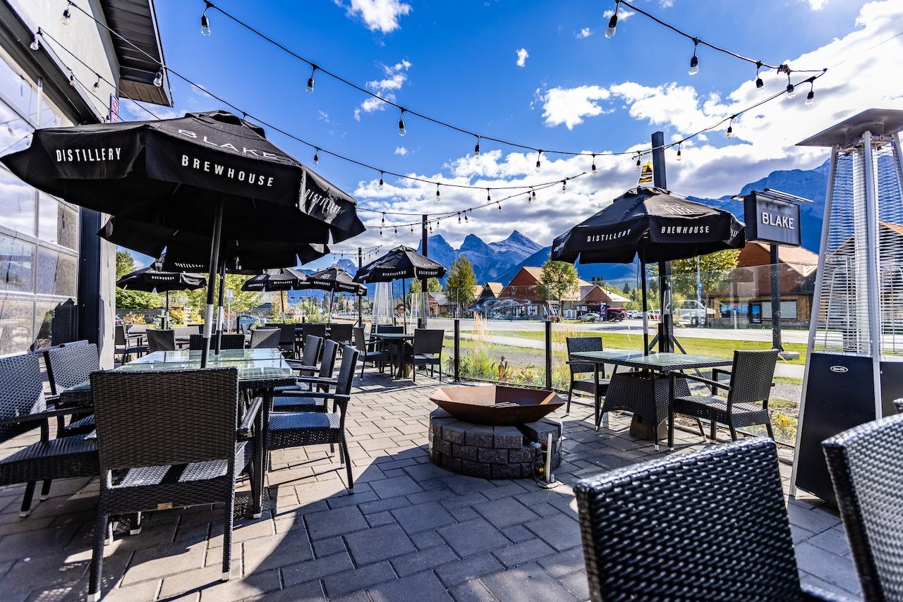 A sunny outdoor patio for Blake Brewhouse Distillery with tables, umbrellas, a fire pit, and mountains in the background.