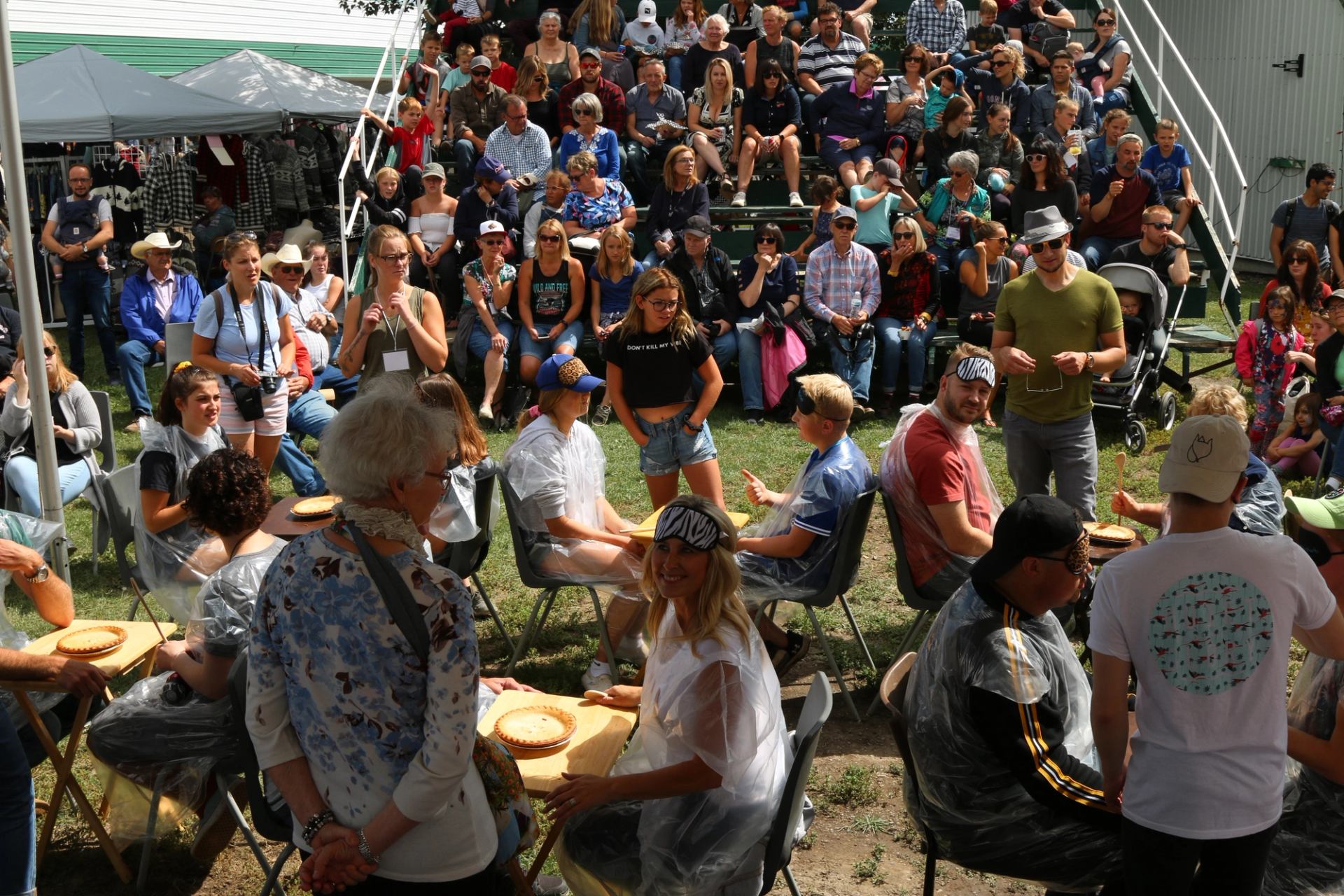 Crowd gathered outdoors around tables and bleachers, watching a lively fair activity on a sunny day.