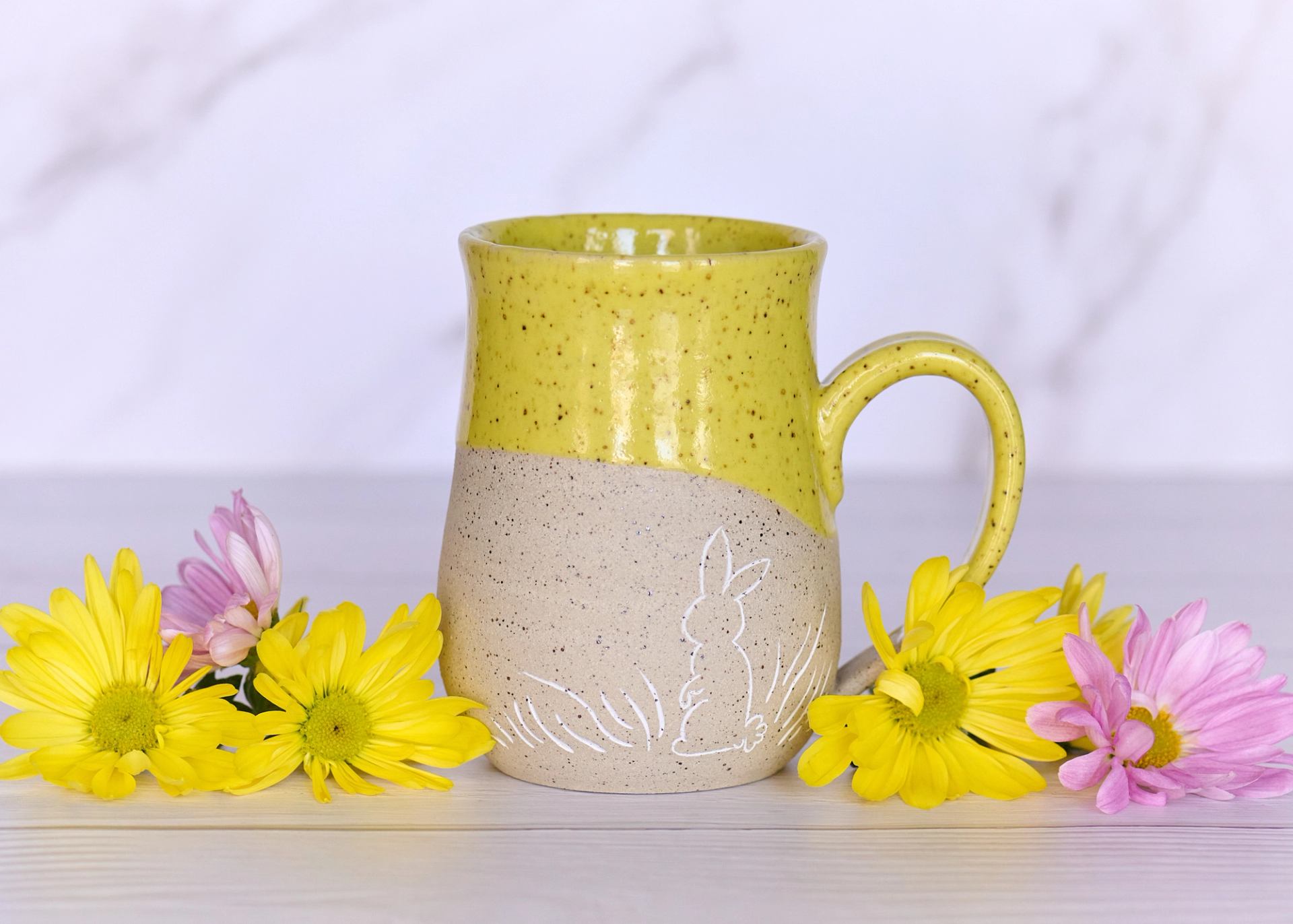 A speckled yellow and beige ceramic mug with a white rabbit drawing, flanked by yellow and pink flowers.