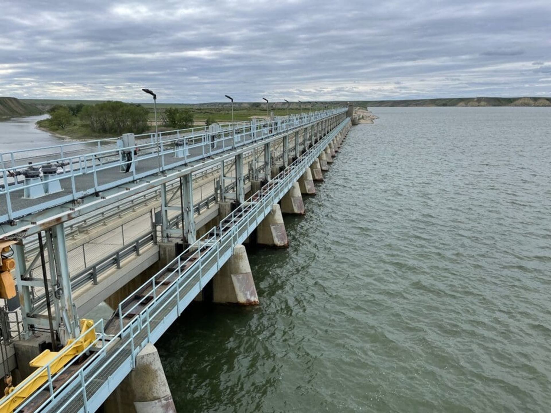 Concrete dam with metal walkways spans wide river under cloudy sky.