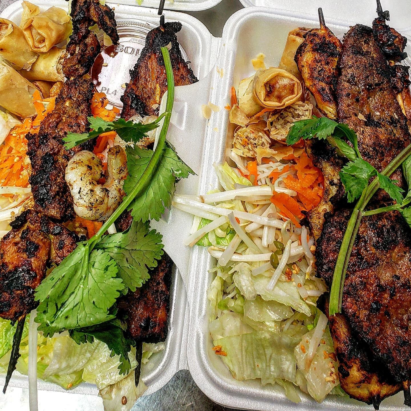 Takeout containers with grilled skewers, veggies, spring rolls, and fresh herbs.