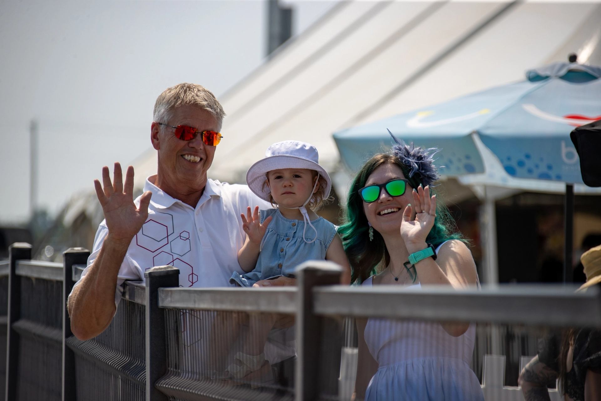 Three people behind a railing wave toward the camera near outdoor tents and umbrellas.