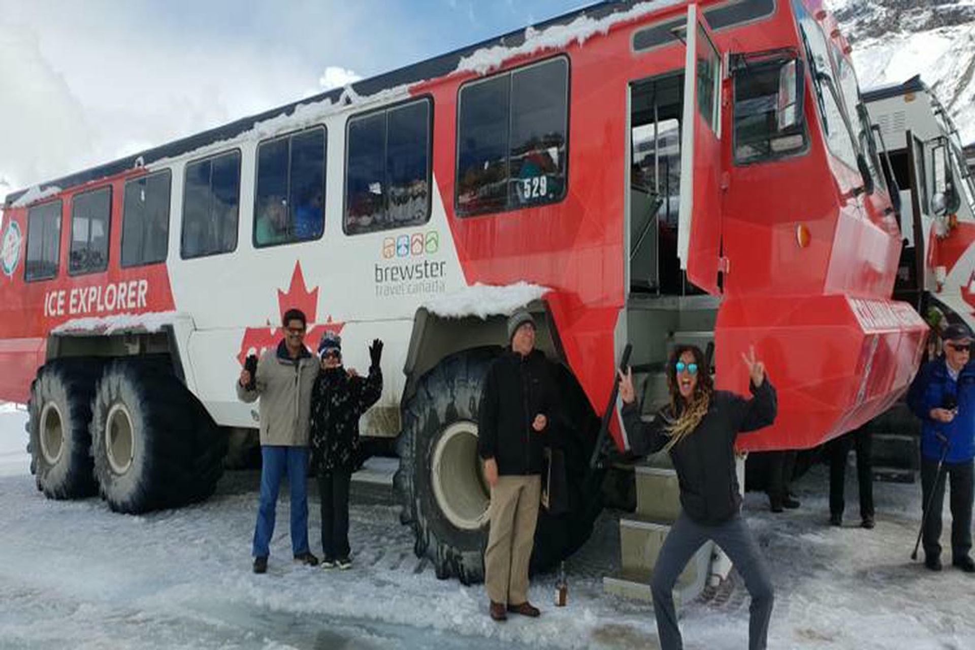 Tourists stand on snow beside red Brewster Ice Explorer bus with large tires.