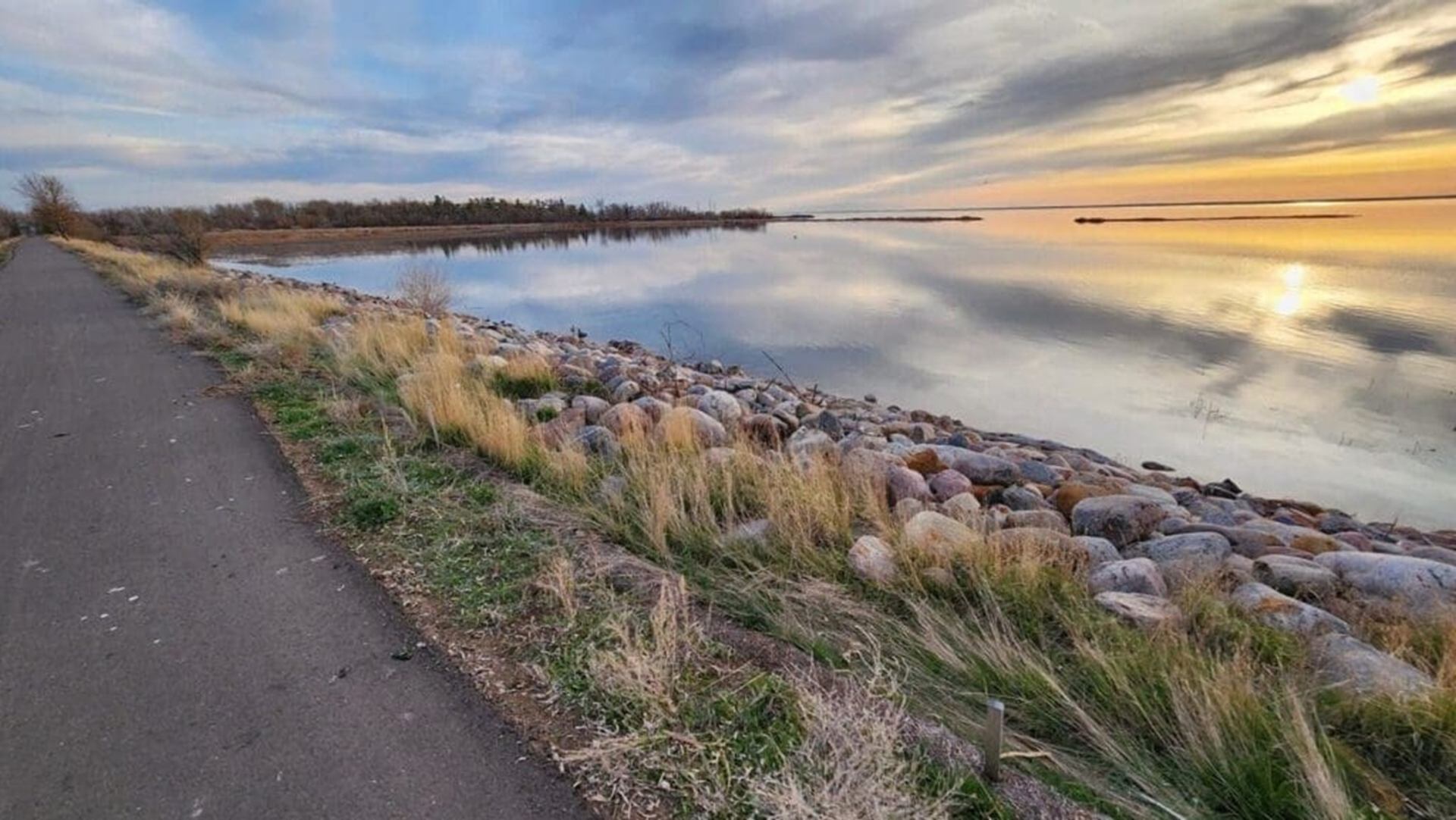 Paved lakeside path beside rocky shoreline with calm water reflecting cloudy sunset sky.